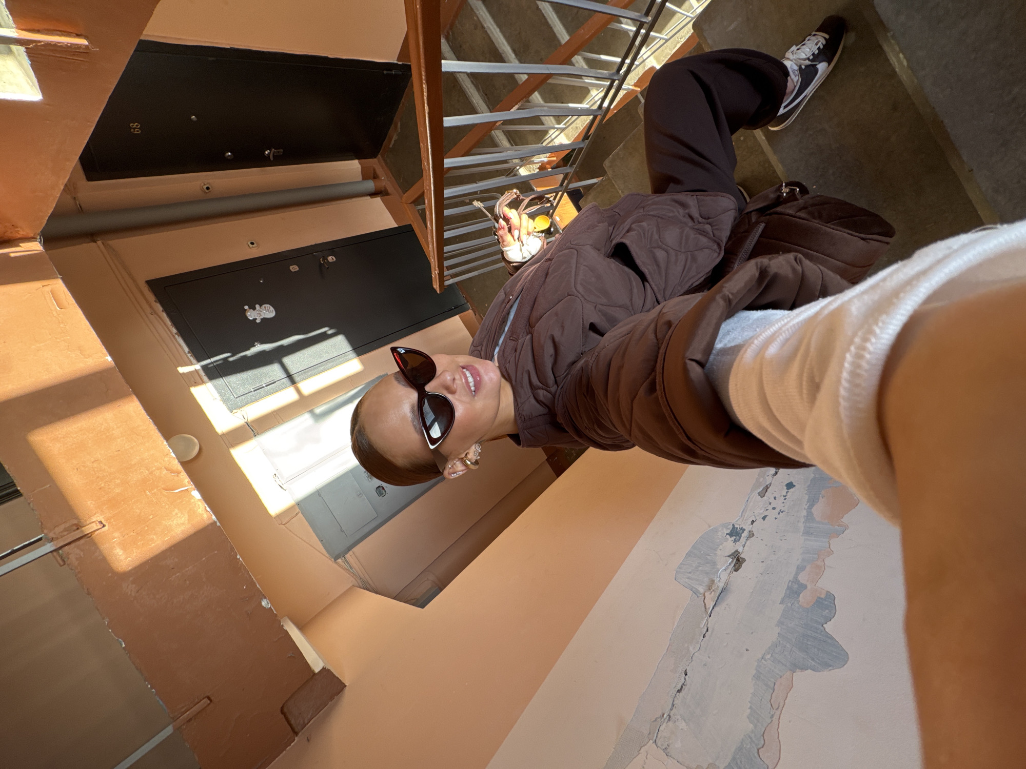 A person wearing sunglasses, a brown jacket, and dark trousers and sneakers stands on a concrete staircase inside a building in what is described to be Chaikovsky, Russia. A brown handbag is slung over their shoulder. The background shows the interior of the building: doors, wall colors, and the staircase structure. The walls are peach-colored with peeling paint. Doors with visible numbering are on the walls. Natural light illuminates the scene.