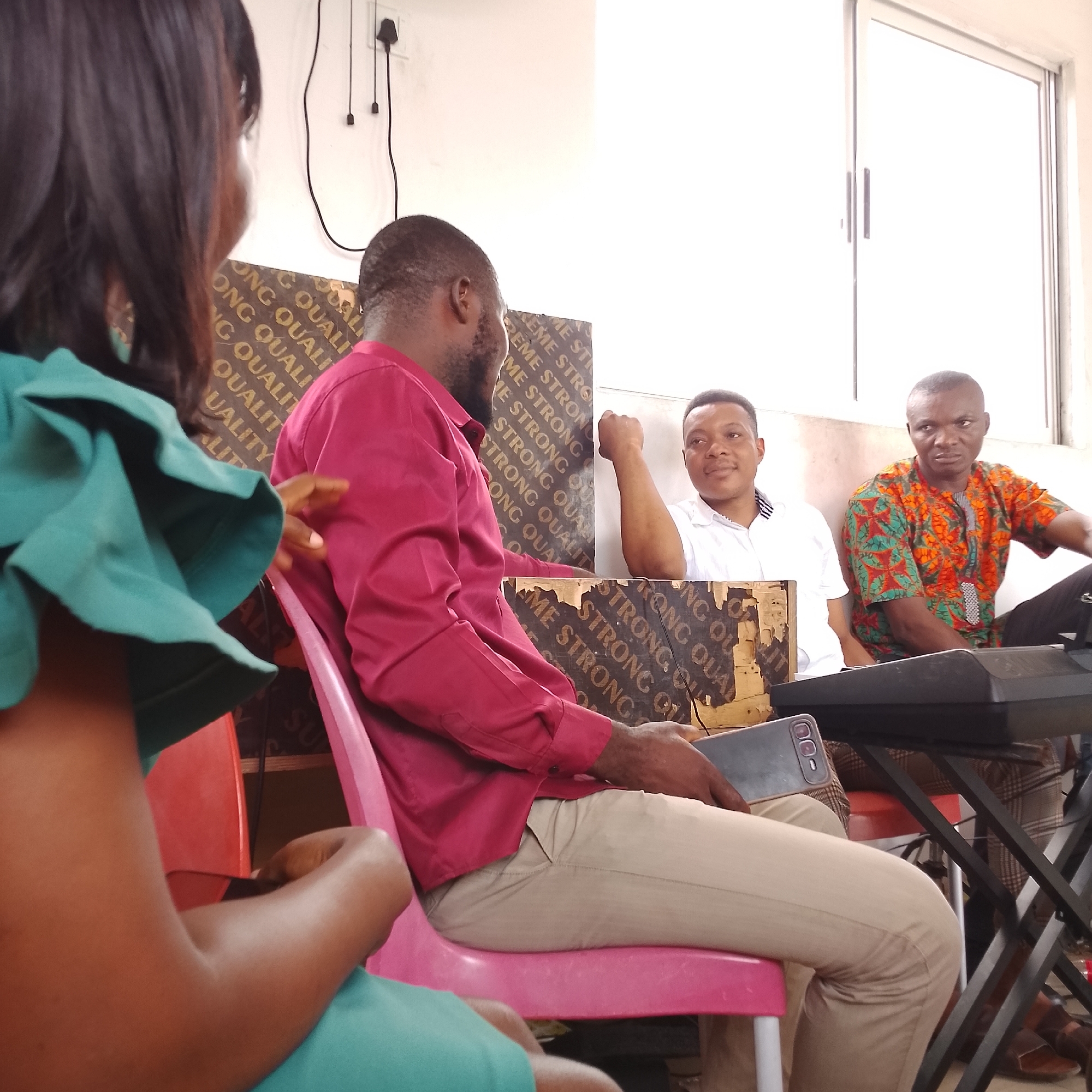 In Iranla, Nigeria, a group of men and one woman are gathered indoors. The woman, partially visible in the foreground, wears a green, ruffled-sleeve dress. A man in a maroon collared shirt and tan pants sits in a pink chair, holding a mobile phone. Another man, wearing a white collared shirt, is seated on the floor with his right arm raised. A fourth man, dressed in a colorful, patterned shirt, is seated to the right. A black keyboard with a metal stand is positioned in front of the man in the patterned shirt. The background features a white wall with electrical outlets and a window, along with wooden boards displaying text. The lighting is bright, suggesting daytime.