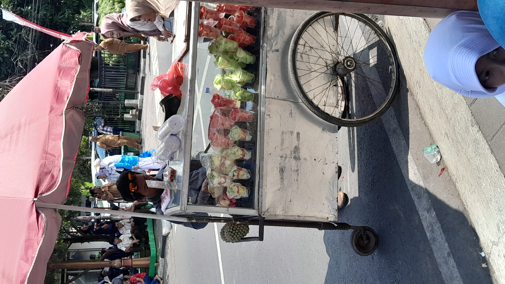 A street vendor in Sumedang, Indonesia, is operating a fruit cart under a large red umbrella. The vendor, a man wearing sunglasses and a dark shirt, is visible behind the glass display of the cart, which is filled with cups of cut fruit. To the left of the cart, a pineapple is attached to a metal rod. In the background, several people are gathered, some appearing to be customers and others possibly onlookers or fellow vendors. The setting appears to be an outdoor urban environment, with a street and sidewalk visible. The cart itself is made of metal and has large wheels, suggesting it is mobile. The overall scene captures a moment of daily commerce in the city.