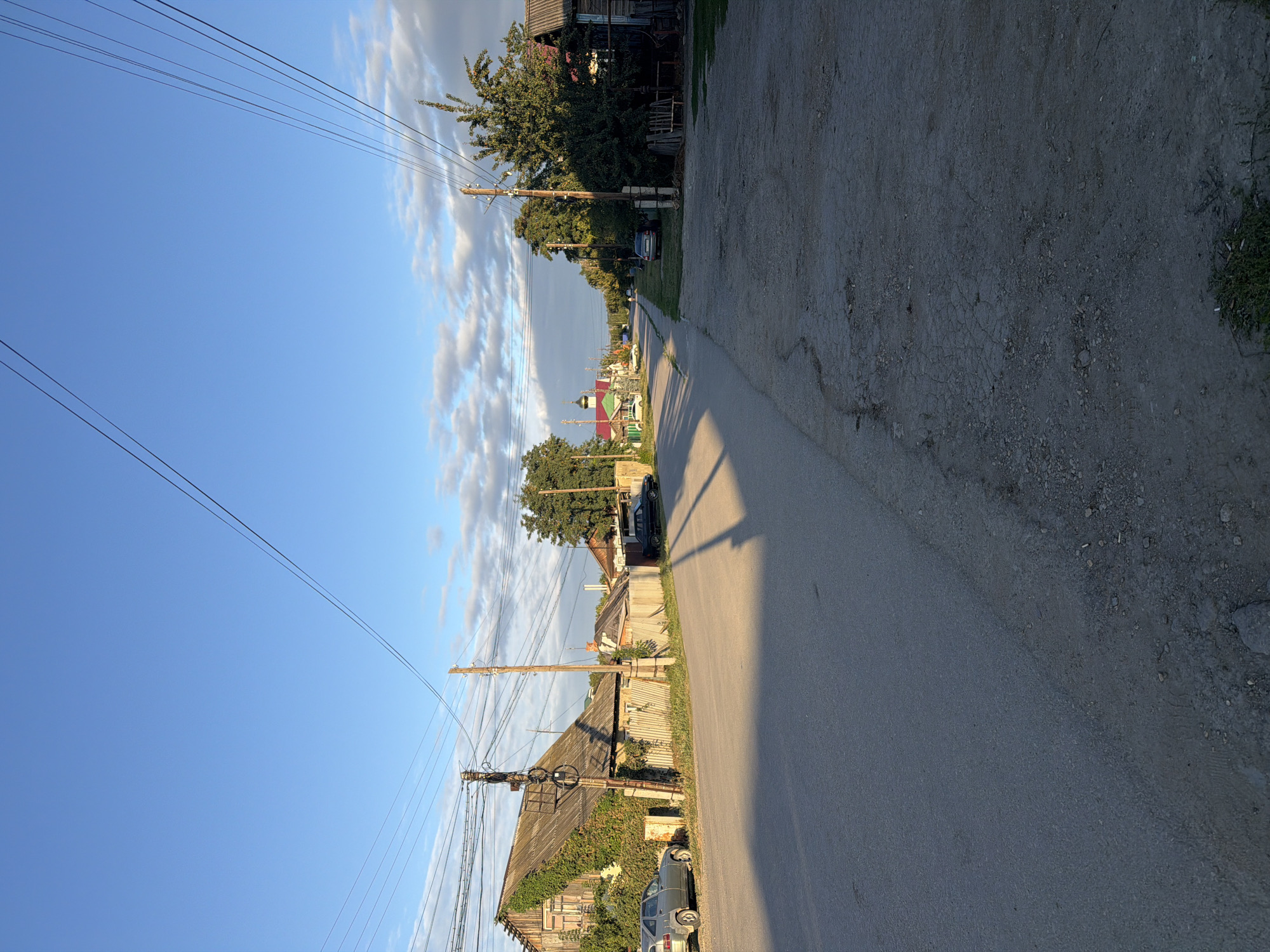 The image shows a street scene in the village of Bagayevka, Russia. The perspective is from the side of the road, looking down a paved street lined with wooden fences and houses. Power lines run overhead, supported by wooden poles. On the left, a grey car is parked in front of a weathered wooden house partially covered in ivy. Further down the street, a dark sedan is parked. In the distance, a church with a green dome is visible. The sky is blue with scattered white clouds, and long shadows suggest it is late afternoon or early evening. The road surface is a mix of asphalt and gravel. There are no people clearly visible in the image.