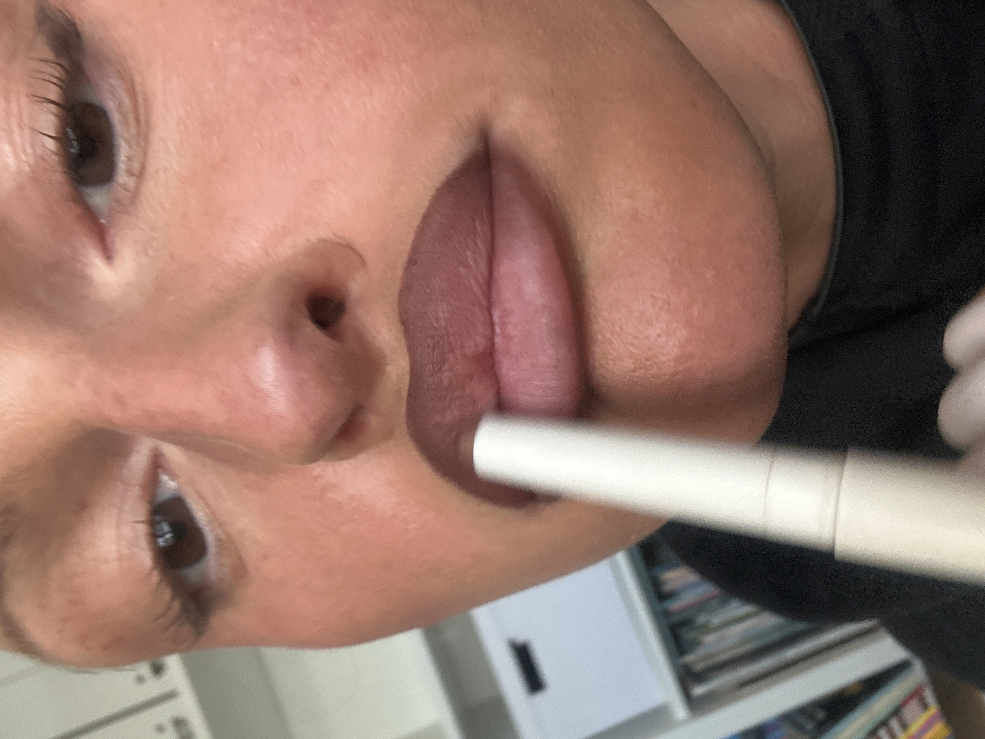The image presents a close-up forensic view of an individual applying a cosmetic product to their lips, located indoors in Wealden, United Kingdom. The subject has medium skin tone, prominent eyelashes, and brown eyes. A white, cylindrical cosmetic applicator is positioned horizontally, making contact with the center of the upper lip. The upper lip displays a partially applied, muted plum-brown pigment, while the lower lip retains a lighter, natural hue. The subject is wearing a dark, possibly black, garment with a visible neckline. The background is blurred, showing a white shelving unit containing a white storage box with a dark rectangular handle and a collection of multi-colored books or binders.