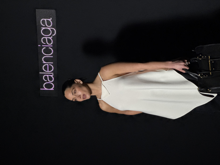 A woman stands in a medium shot against a plain black background. She has dark hair pulled back from her face and wears a sleeveless, asymmetrical off-white top with thin straps and a distinct V-neckline detail. Her gaze is directed to her left, with her mouth slightly open. In her right hand, she holds a structured black handbag with visible gold-colored hardware. Above and behind her, centered in the upper portion of the frame, a luminous magenta LED sign displays the word "balenciaga." A faint shadow of the woman is cast on the black surface to her right. The scene likely occurs at a Balenciaga event, consistent with the brand's presentations often held in Paris, France.