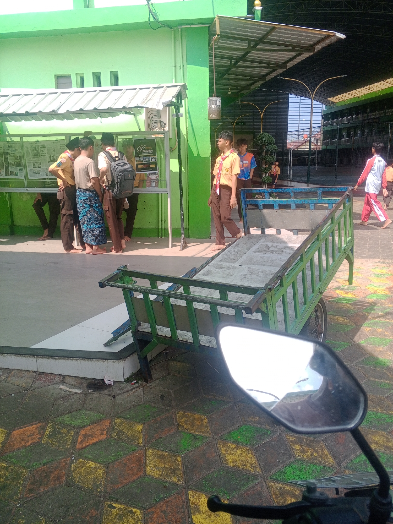 In Ponorogo, Indonesia, a group of young students in uniform, likely from a school, are gathered outside a building with a bright green facade. Some are wearing tan shirts and brown pants, characteristic of scouting uniforms, and appear to be looking at a display board. One student is wearing a patterned sarong over his pants. In the foreground, a green and blue cart with a worn wooden bed is parked on a sidewalk with a distinctive patterned pavement of interlocking cubes in shades of green, yellow, and orange. The students' bare feet are visible. In the background, a covered walkway leads to a sports court, and several other people, also in uniforms, are walking. The overall scene suggests a school environment during daytime, possibly in the morning or afternoon, given the bright sunlight.