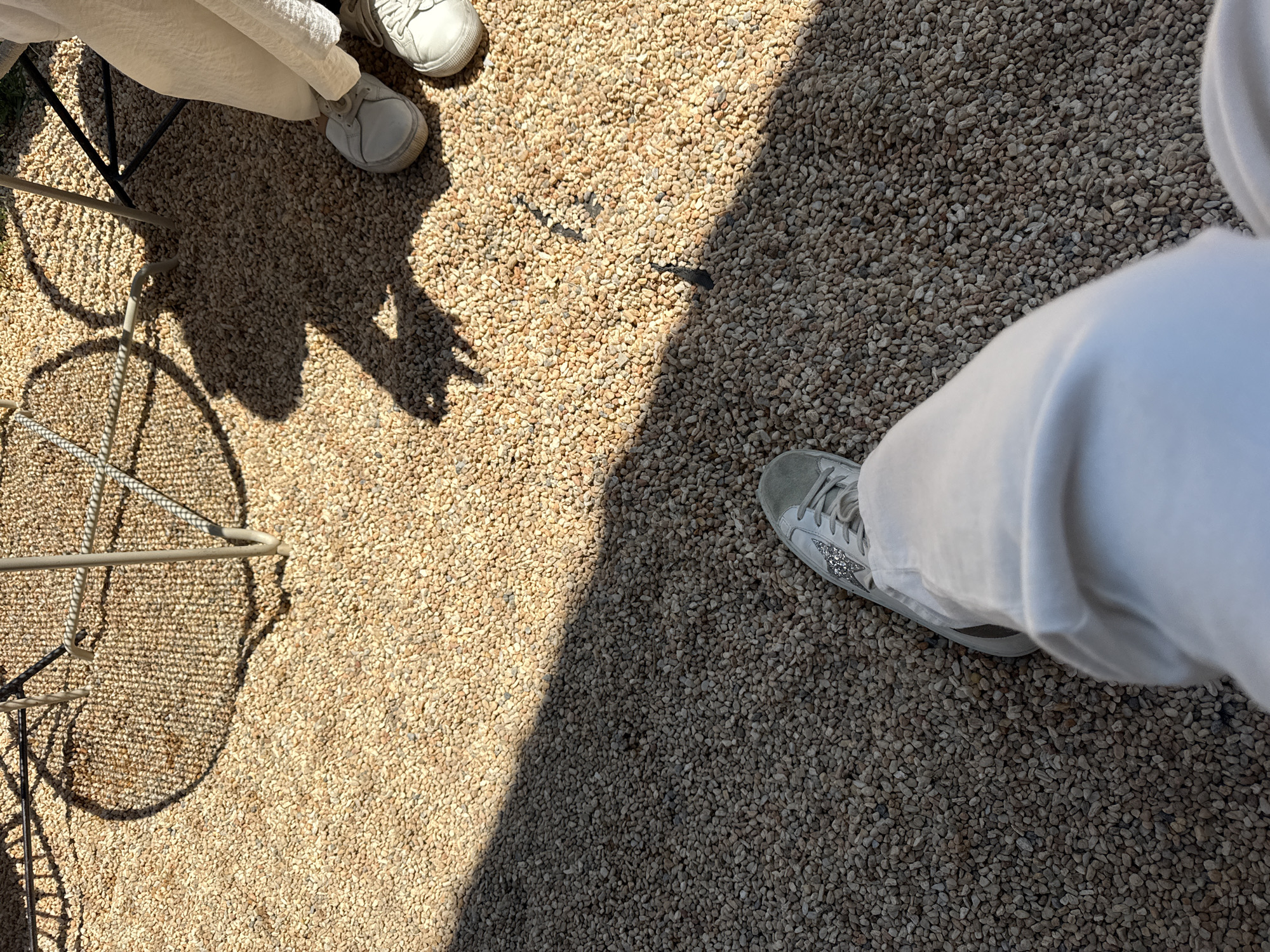 The image shows a ground-level view of a gravel surface in San Andrés Cholula, Mexico. The scene is partially lit by sunlight, creating a distinct shadow dividing the frame. In the upper left corner, there are two pairs of white sneakers. A metal chair leg with a circular mesh seat is also visible, casting a shadow on the gravel. In the lower right corner, part of a white outfit is visible along with a single sneaker. The gravel consists of small, closely packed pebbles.