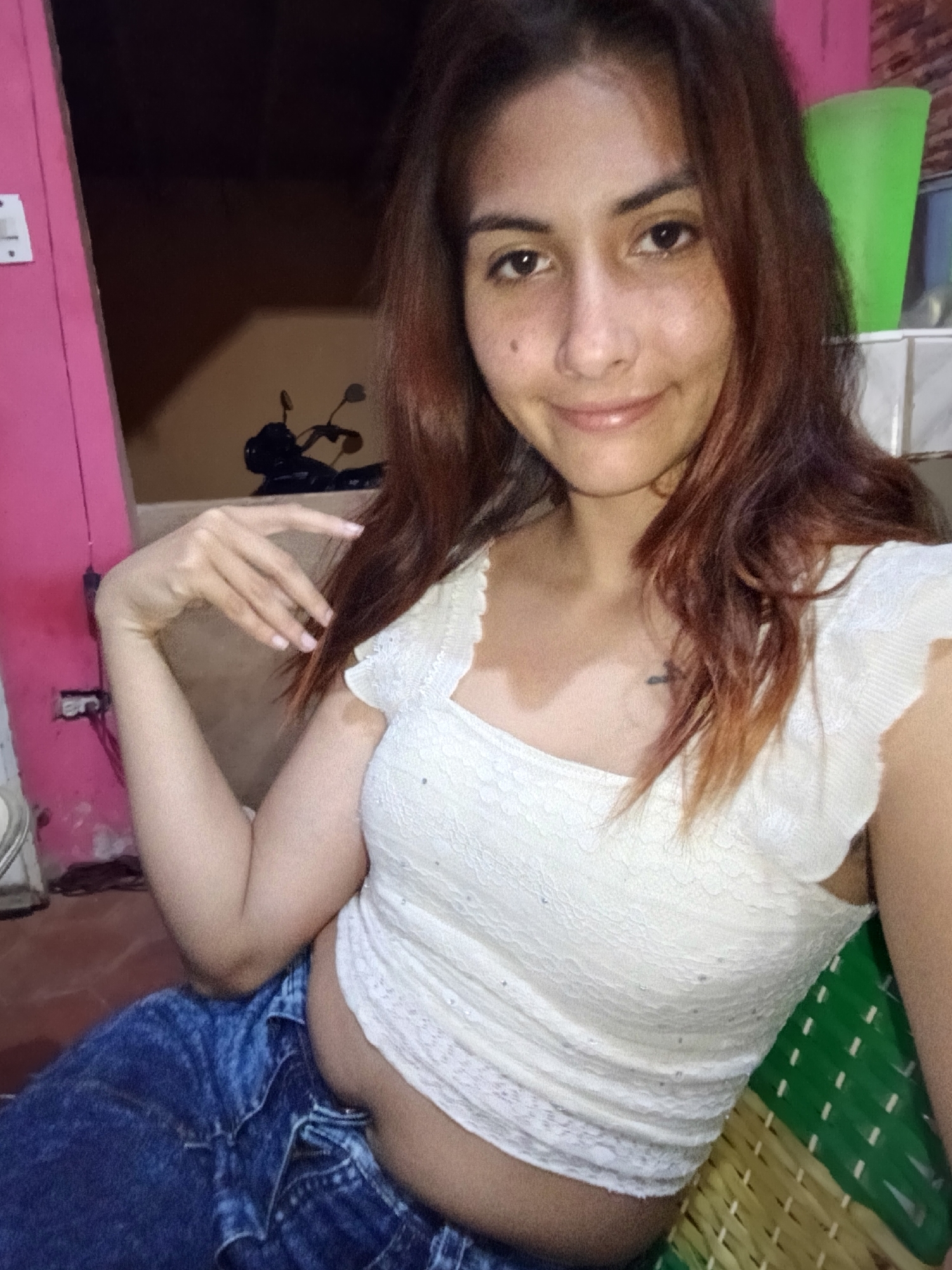 The image shows a young woman with reddish-brown hair and light skin, wearing a white lace top and blue jeans. She is seated, with her left arm bent and hand resting near her head, and her right arm is also bent. Her gaze is directed towards the camera with a slight smile. In the background, there is a pink wall on the left, and a motorcycle is partially visible. To the right, a green plastic cup and a woven mat are present. The setting appears to be indoors or a covered outdoor area. The overall lighting suggests it is nighttime or a dimly lit environment. The image does not provide any specific visual cues to confirm the location in Juan Griego, Venezuela.