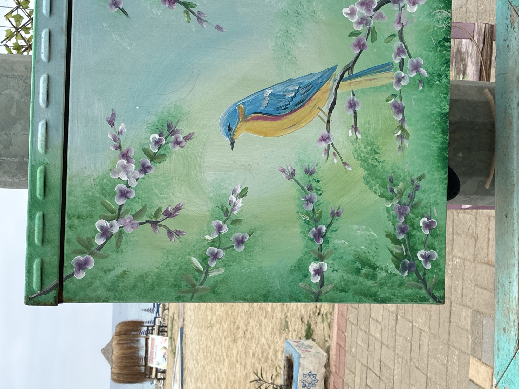 The image shows a painted metal utility box on a beach in Alanya, Turkey. The box is painted with a green background and features a painting of a blue and yellow bird perched on a branch with purple flowers. In the background, there is a sandy beach with lounge chairs and a thatched umbrella. The sky is overcast. The utility box is located outdoors, likely in a public space near the beach.