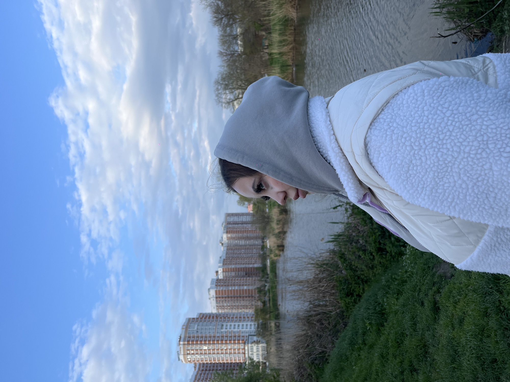 A young woman stands in profile on the right side of the frame, looking downwards and to her left. She is wearing a grey hooded sweatshirt underneath a cream or off-white quilted vest with a textured, fluffy sherpa lining. Her dark hair is pulled back, and she has visible eyelashes.
The scene is set outdoors along the bank of a body of water, likely a river or canal, in Krasnodar, Russia. The water surface is calm, reflecting the cloudy sky. On the far bank of the waterway, a series of tall, modern residential buildings with numerous windows and balconies stretch into the distance, indicating an urban environment. Some trees, appearing to be bare, are visible both near the buildings and further along the river. The immediate foreground features lush green grass and bushes.
The sky above is a mix of clear blue patches and soft, white clouds, suggesting it is daytime with diffused light. The weather appears cool, as indicated by the woman's warm attire. There is no specific activity taking place other than the woman observing her surroundings. No text is visible in the image.