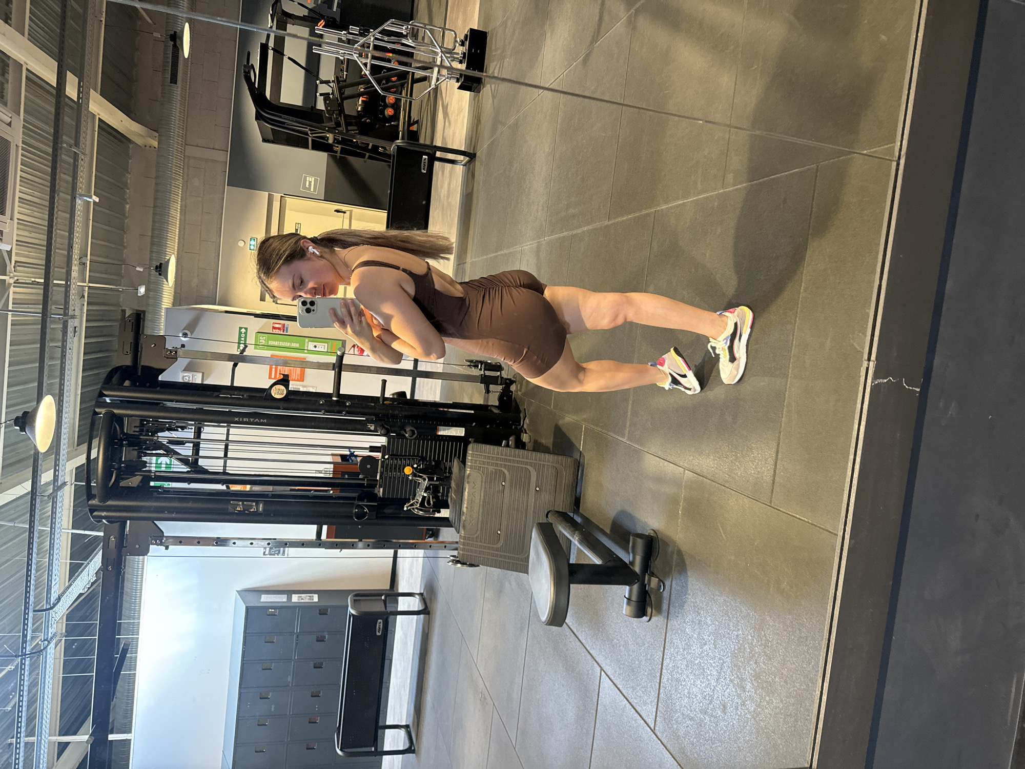 A woman is taking a mirror selfie in a gym. She is wearing a brown, sleeveless one-piece outfit and multi-colored sneakers. She has her hair pulled back in a ponytail and is wearing earbuds. The gym has grey tiled flooring and various exercise machines, including a cable machine, a weight rack, and a bench. In the background, there are lockers and industrial ceiling elements with lighting. The reflection shows the woman from a three-quarter view. The scene is clean and well-lit, typical of a commercial gym environment. There is no information in the image that suggests the location is the city of Saint-Claude, France.