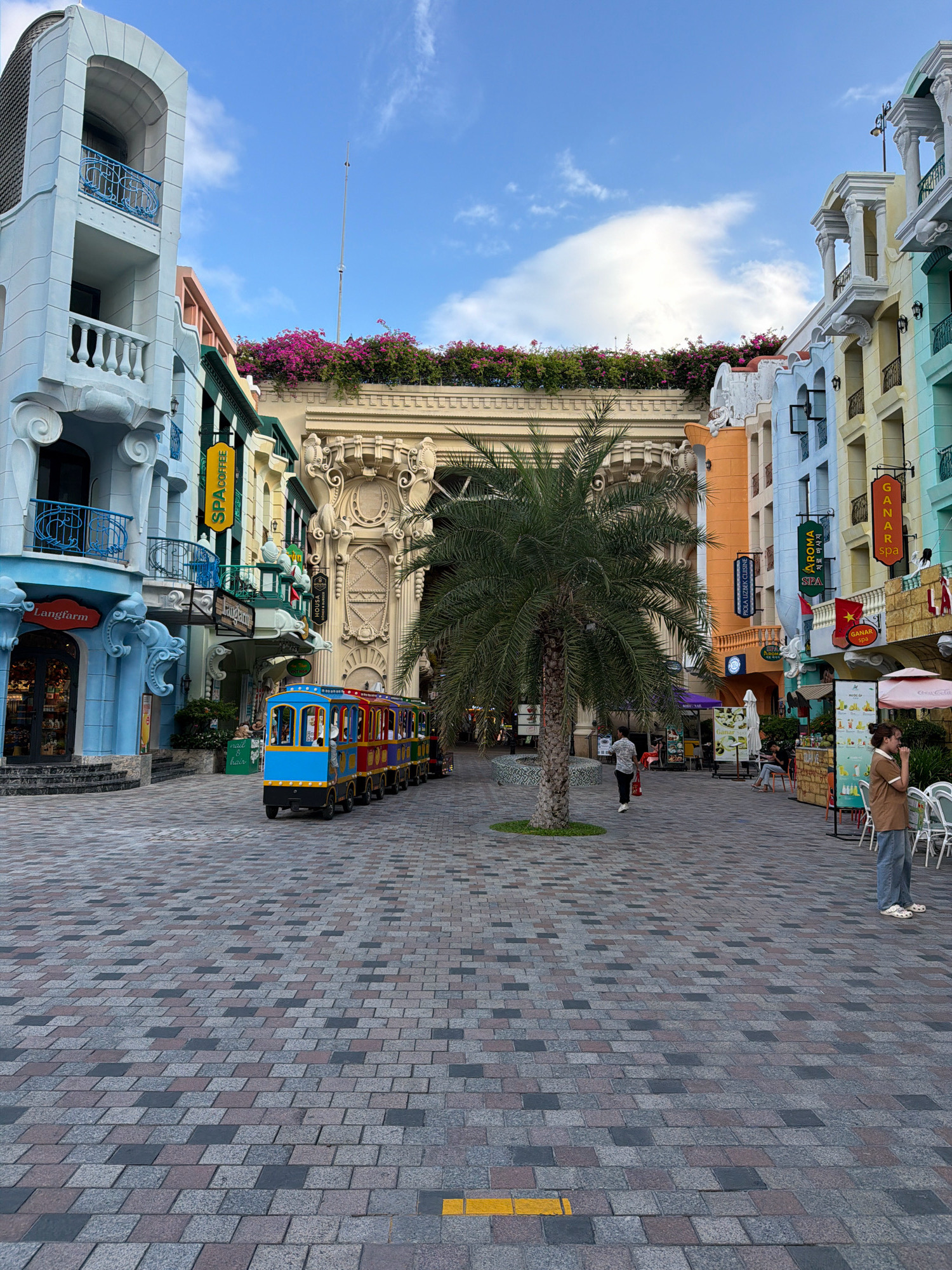 The image depicts a street scene in Nha Trang, Vietnam, likely within a tourist-oriented area, possibly a resort or entertainment complex. The architecture is a vibrant mix of colors and styles, reminiscent of European influences, with pastel-colored buildings featuring balconies, columns, and ornate detailing. A small, multi-colored train, resembling a miniature amusement park ride, travels down the cobblestone street. A palm tree stands prominently in the foreground. Several people are visible: a woman in a brown top and jeans stands near tables and chairs, another woman sits at a table on the right, and a person walks away from the camera towards the train on the left. The sky is blue with scattered clouds. The ground is paved with patterned cobblestones. Signs for businesses such as "Langfarm," "SPA coffee," "AROMA SPA," and "GANAR SPA" are visible on the buildings, indicating commercial activity.