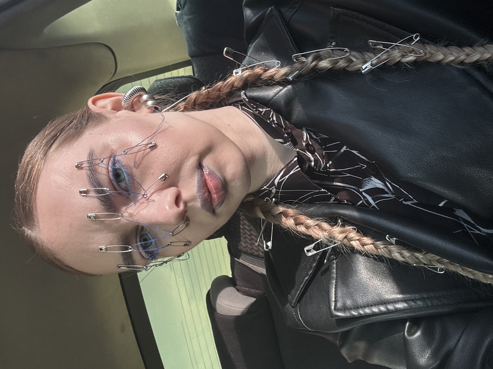 The image shows a woman's face and upper body, seemingly inside a vehicle. She has fair skin and blonde braided hair adorned with several silver safety pins. Similar safety pins are attached to her face, connected by thin blue string around her eyes and nose. She is wearing blue eye makeup and has a dark lipstick color with blue undertones. The woman is wearing a black leather jacket over a black and white patterned top. The background shows the interior of a car, with a green door panel visible in the lower left corner. The location is unspecified as Krasnoyarsk, Russia. The angle of the shot is slightly tilted.