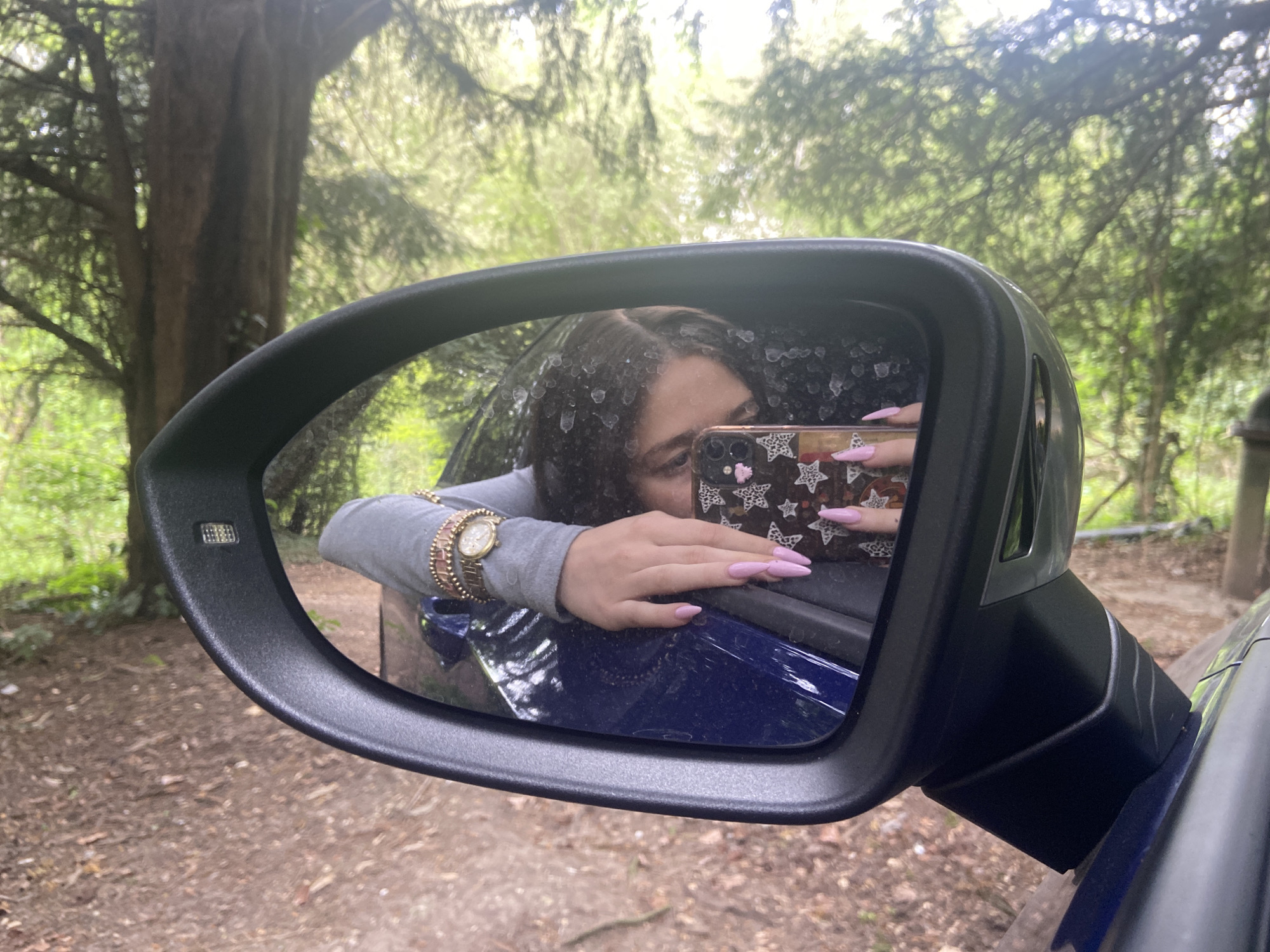 An outdoor, eye-level shot captures a person taking a selfie reflected in the side mirror of a blue car, set against a verdant, wooded backdrop in Upper Basildon, United Kingdom. The black-framed car mirror, positioned centrally, shows the upper body of a person, likely female, leaning against the car's window frame.
The person is wearing a grey long-sleeved top and has long, light pink, almond-shaped nails. A gold-toned watch with a round face and multiple gold-toned bracelets adorn their left wrist. Their right hand holds a smartphone, partially obscuring the face, which has a black and white patterned case featuring stars or leopard print. The car's exterior, visible beneath the mirror, is a deep blue. The mirror surface exhibits numerous water spots or residue.
The background, visible through and around the mirror, is dominated by tall trees with lush green foliage, suggesting a forest or dense woodland. The ground visible beneath the car is an unpaved dirt path, covered with fallen leaves and debris. The natural lighting indicates it is daytime, with light filtering through the tree canopy. The scene suggests a casual moment captured during an outdoor excursion or a stop in a scenic natural environment. No visible text is present in the image.