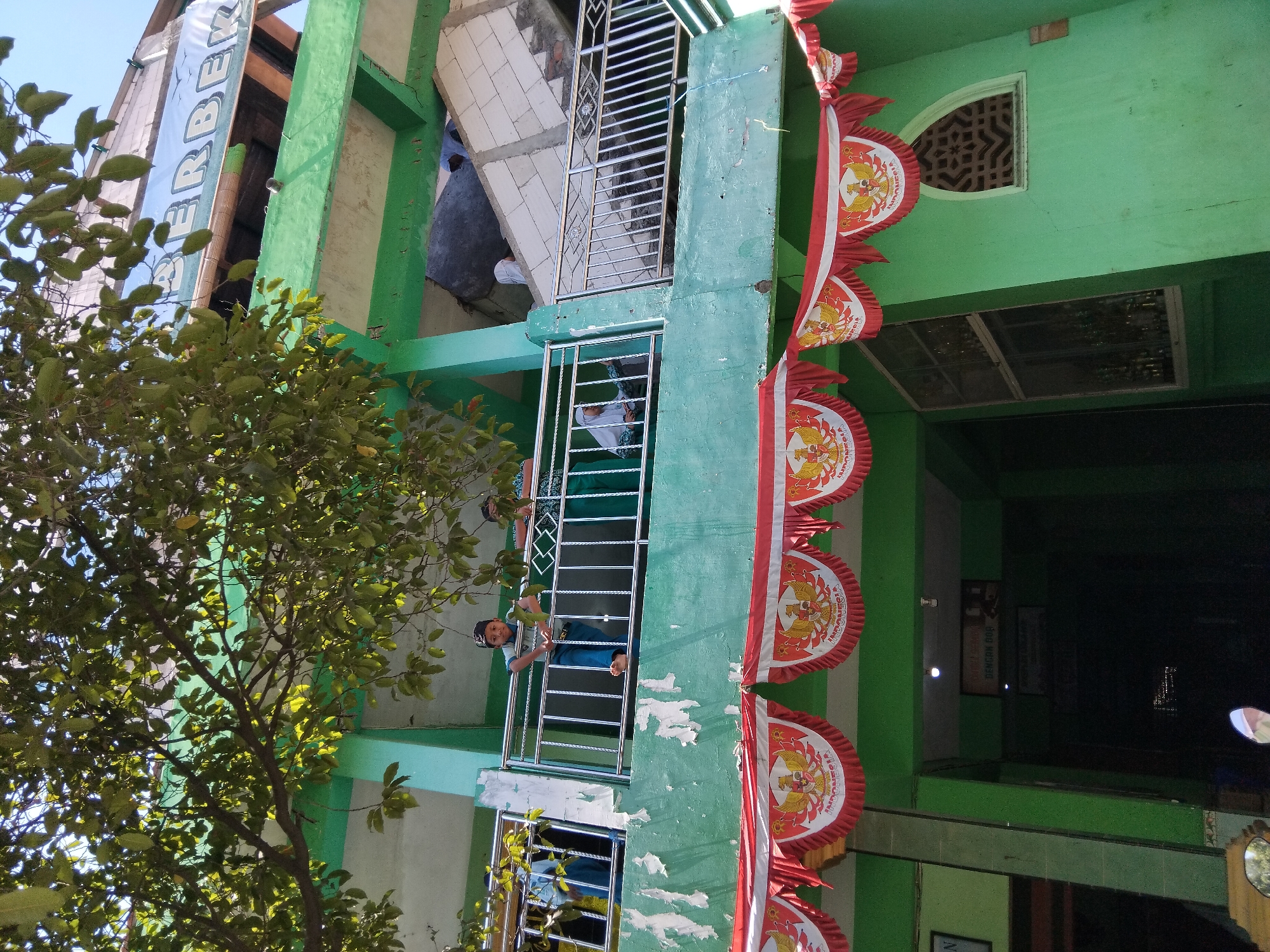 The image depicts a two-story green building, likely a school or community center, in Kepuh Kiriman, Indonesia. The lower level features a dark entrance with signs and a decorative arched window. A prominent red and white banner, featuring the Indonesian emblem, is strung across the front of the building, suggesting a national celebration. The upper level has a balcony with a metal railing, where several individuals are visible. On the left, a person in a blue shirt and pants stands, while to the right, two individuals, one wearing a white hijab and the other a patterned green garment, are also present. A pile of construction debris, including concrete blocks and rebar, is visible on the upper floor, indicating ongoing or recent construction or damage. Lush green foliage from a large tree overhangs the top of the building, casting shadows and partially obscuring a banner with the word "BERBEK" visible at the top. The sky is clear and blue.