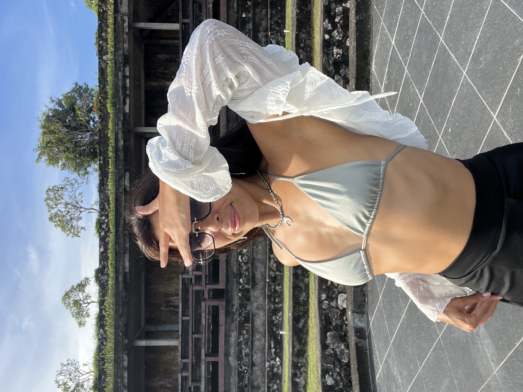 A woman poses outdoors under a clear blue sky, likely in Kuta Selatan, Indonesia. She is depicted from the waist up, wearing a light grey or silver bikini top overlaid with a sheer white lace long-sleeved shirt, and dark bottoms. Sunglasses are on her face, and a silver necklace with a unique 'C' or crescent-shaped pendant is visible. Her arms are raised, one hand near her head, as she looks towards the camera with a subtle smile, conveying a relaxed and confident demeanor.
The background features weathered stone structures, possibly steps or walls of an ancient site, partially covered with green moss and vegetation. Several tropical trees with sparse foliage stand against the bright sky. The ground where she stands is paved with dark grey tiles, forming a diamond-grid pattern with white grout lines. Strong sunlight suggests it is daytime, casting clear shadows. No visible text is present in the image.