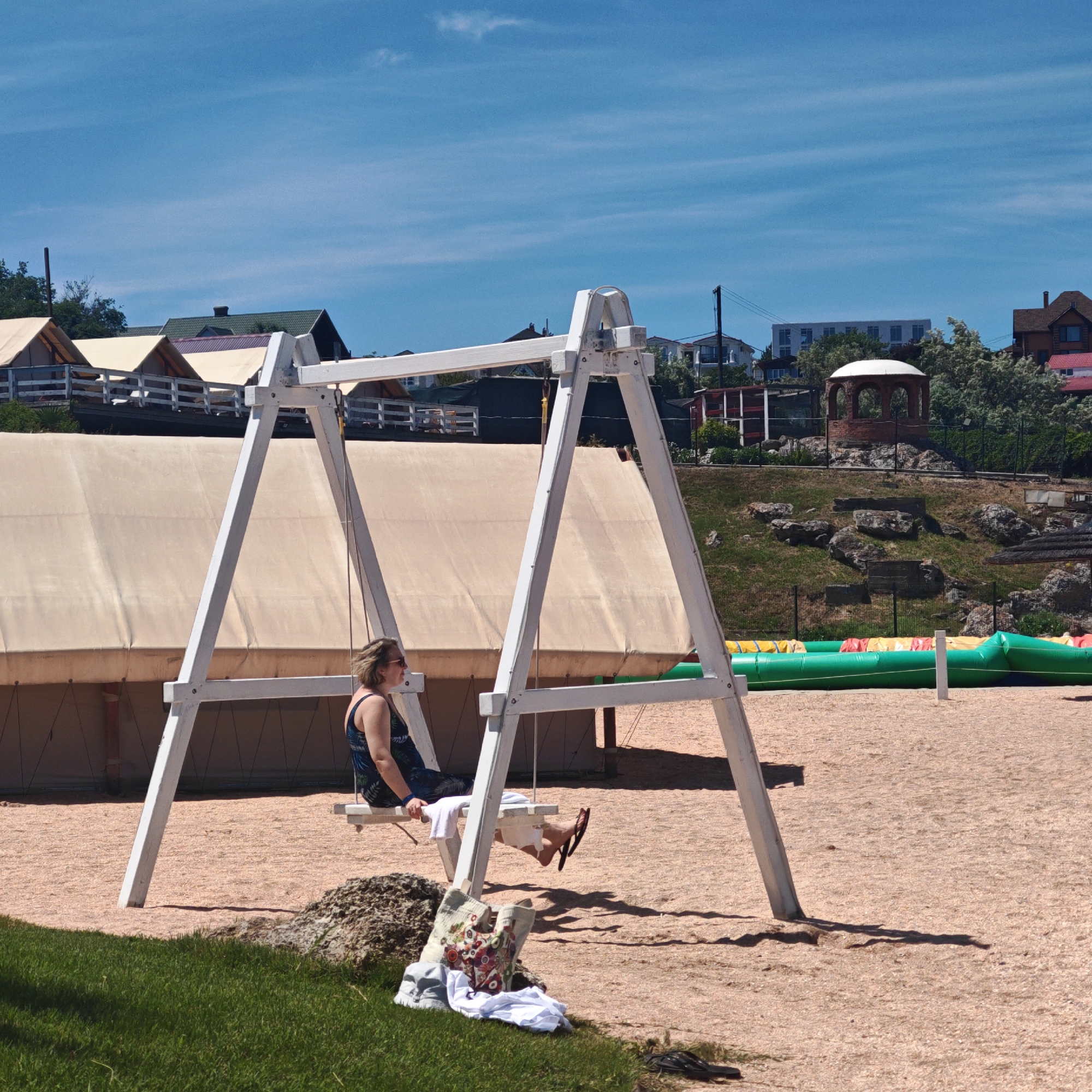 The image displays an outdoor leisure area under a bright, clear blue sky with minimal cirrus clouds. In the foreground, a patch of green grass is visible on the left, transitioning into an extensive area of light brown, granular ground, possibly wood chips or fine gravel. A prominent white wooden A-frame swing set is positioned centrally. An adult female, appearing Caucasian with light-colored hair and wearing sunglasses, a dark-patterned sleeveless top (possibly a swimsuit or sundress), and black flip-flops, is seated on the right swing. She faces left, with her right leg bent and her left leg extended. A white towel is draped on the swing seat adjacent to her. Near the bottom left, beside the grass and a dark rock, a light-colored tote bag with a multi-colored abstract pattern, a grey cap, and white folded clothing are placed on the ground.
In the midground, a large, light-beige canvas tent structure is situated behind the swing set. Above this, on a raised dark platform with white wooden railings, several smaller tent-like structures with light-colored roofs are visible. The background consists of a green hillside dotted with rocks and sparse trees. A brick gazebo with a white domed roof is centrally located on this hillside. Various buildings, including residential structures with dark green and red roofs, are visible on the distant hilltop. To the far right, on the light brown ground, multiple long, green inflatable structures with yellow and red segments are present, suggesting recreational equipment. Strong shadows cast by the swing set indicate direct sunlight. The scene depicts a sunny, outdoor recreational or resort environment.