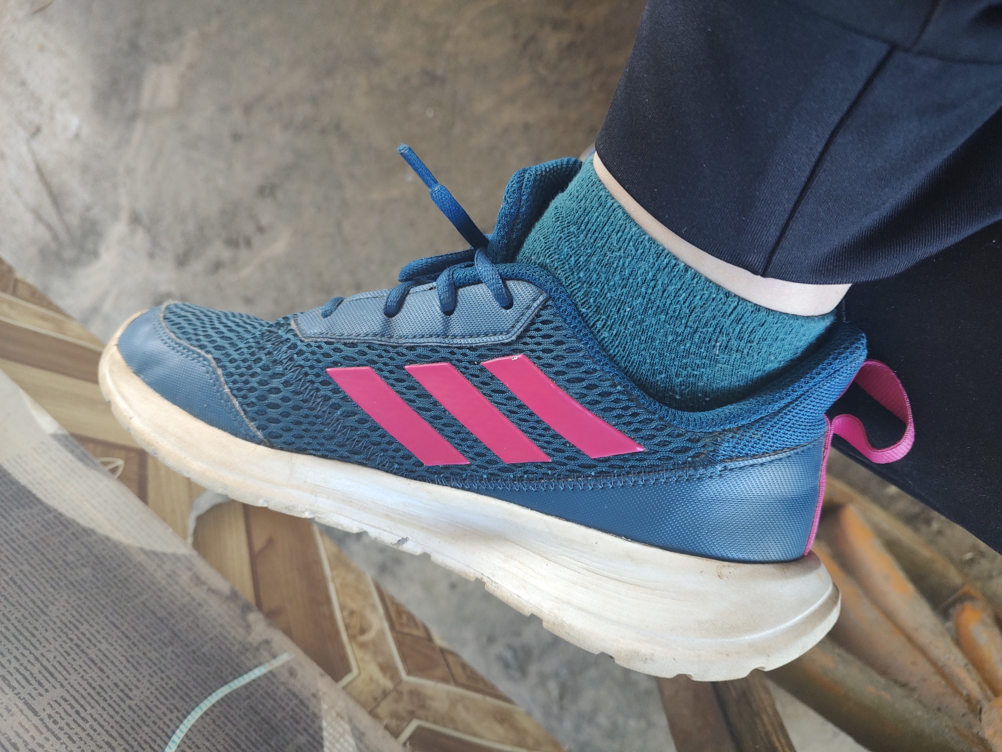 In Kochmar, Bulgaria, a close-up, high-angle shot captures a person's left foot wearing a blue and pink Adidas athletic shoe. The shoe features a blue mesh upper with three prominent pink stripes on the side. A blue lace is visible, loosely tied. The wearer is also wearing a teal colored sock and black pants. The shoe rests on a surface composed of what appears to be a weathered wooden bench and a concrete floor. The lighting suggests an outdoor or semi-outdoor environment, possibly during daylight hours. The overall impression is of a casual, everyday setting.