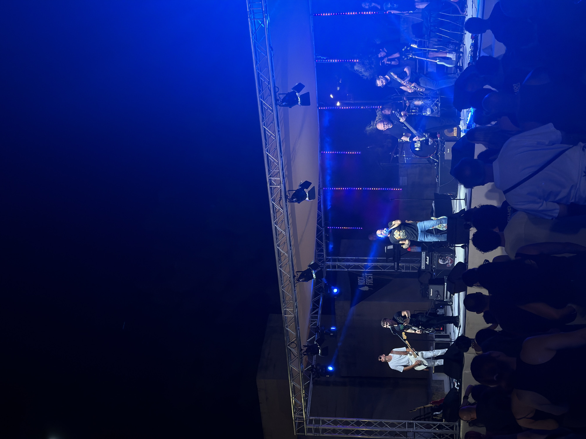 The image depicts an outdoor music concert at night in Saint Paul's Bay, Malta. A band performs on a stage illuminated by bright blue lights. The band consists of several musicians, including guitarists, a vocalist, and a drummer. They are all dressed in casual attire and appear to be actively engaged in their performance. The stage is set up with musical instruments, amplifiers, and lighting equipment. In front of the stage, a crowd of spectators is gathered, silhouetted against the bright stage lights. The background reveals a dark night sky and some architectural elements, possibly buildings or structures common in Saint Paul's Bay. The overall atmosphere is energetic and festive, indicative of a live music event.