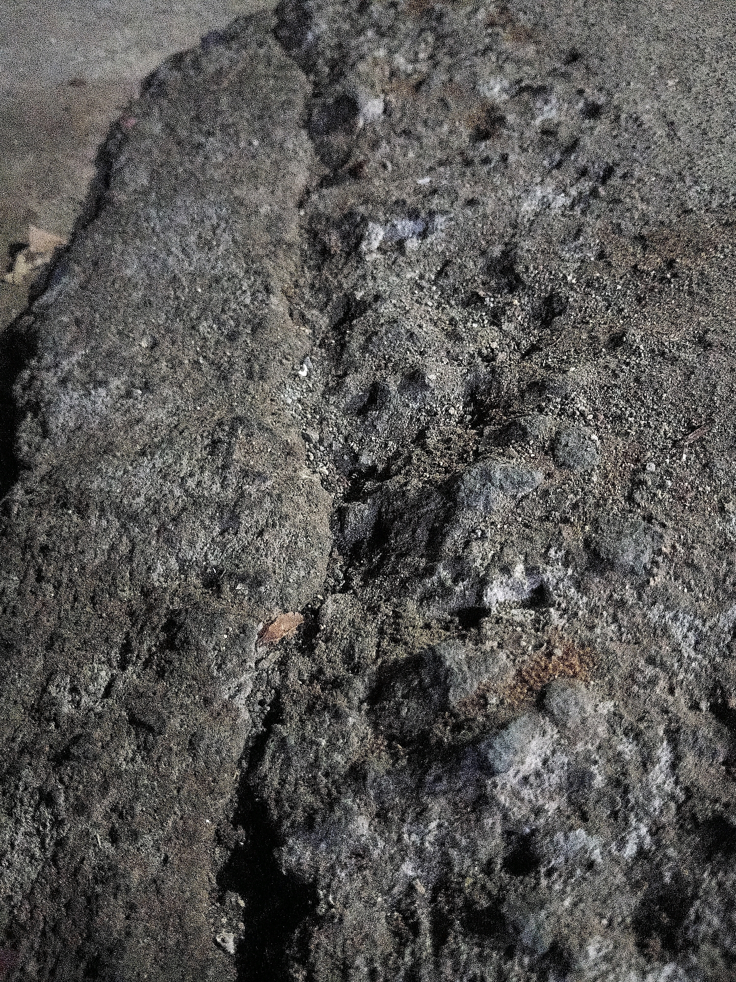 The image shows a close-up view of a cracked and weathered concrete surface, possibly part of a sidewalk or road. The concrete is dark gray and rough in texture, with visible aggregate and debris embedded in it. A prominent crack runs diagonally across the image, separating two sections of concrete that appear to have shifted slightly. The surrounding area is also concrete, with some areas appearing lighter and possibly dusty. There are no people, animals, or specific landmarks visible in the image to indicate a precise location in Cikal, Indonesia. The scene is static and appears to be an outdoor environment.