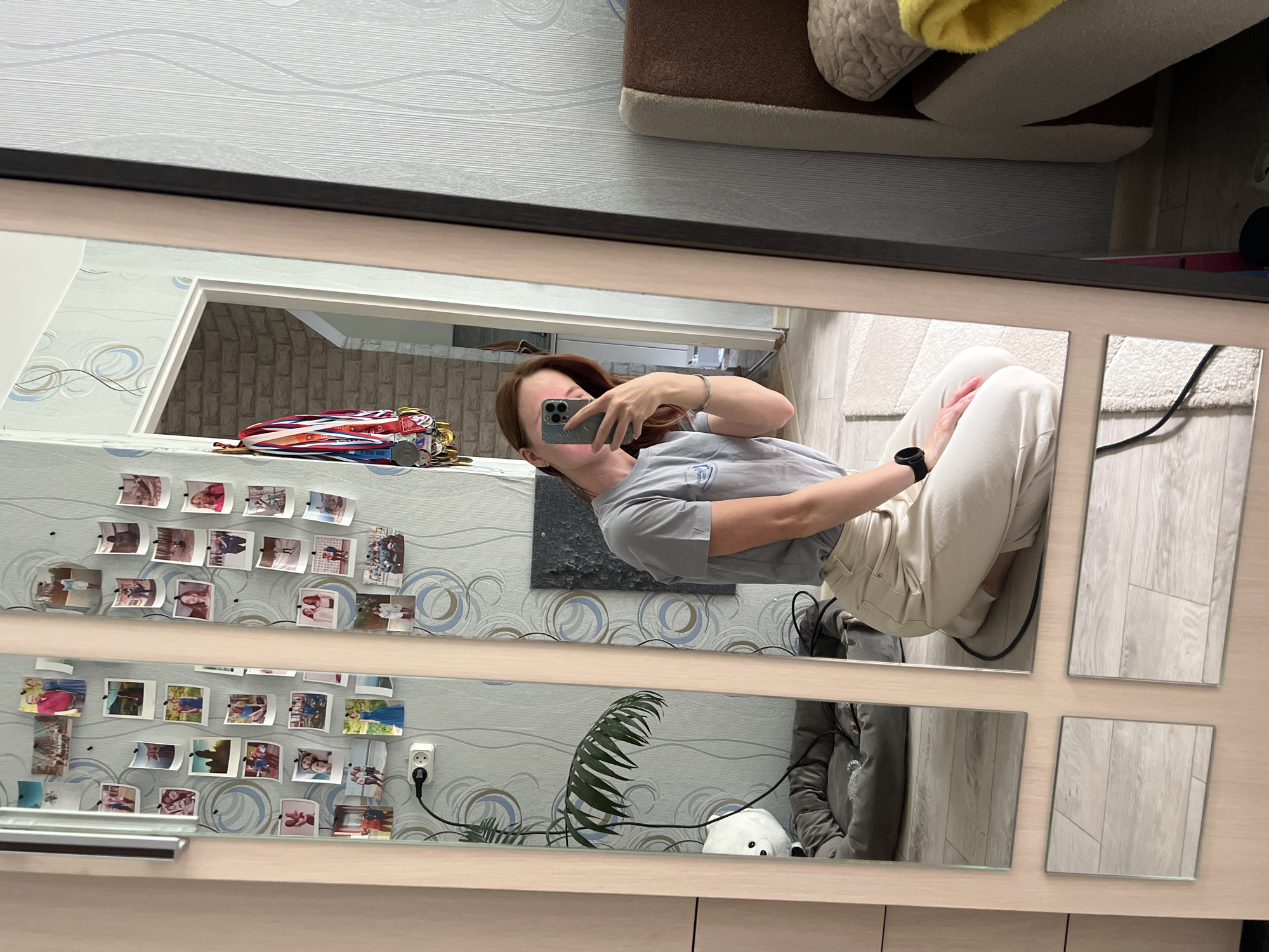 In a bedroom, a young woman with brown hair is kneeling on the floor, taking a selfie in a full-length mirror. She is wearing a gray t-shirt and cream-colored pants. Her phone is held up to capture her reflection. The mirror is attached to a closet door, and the reflections show a wall decorated with numerous small photographs and a few medals hanging on a hook. A houseplant is visible in the reflection of the mirror, near a dog bed with a white fluffy toy on it. The room is lit by natural light. The image shows an interior setting that could be a bedroom. The provided context states the location is the city of Chaikovsky, Russia, though this is not visually evident from the image itself.