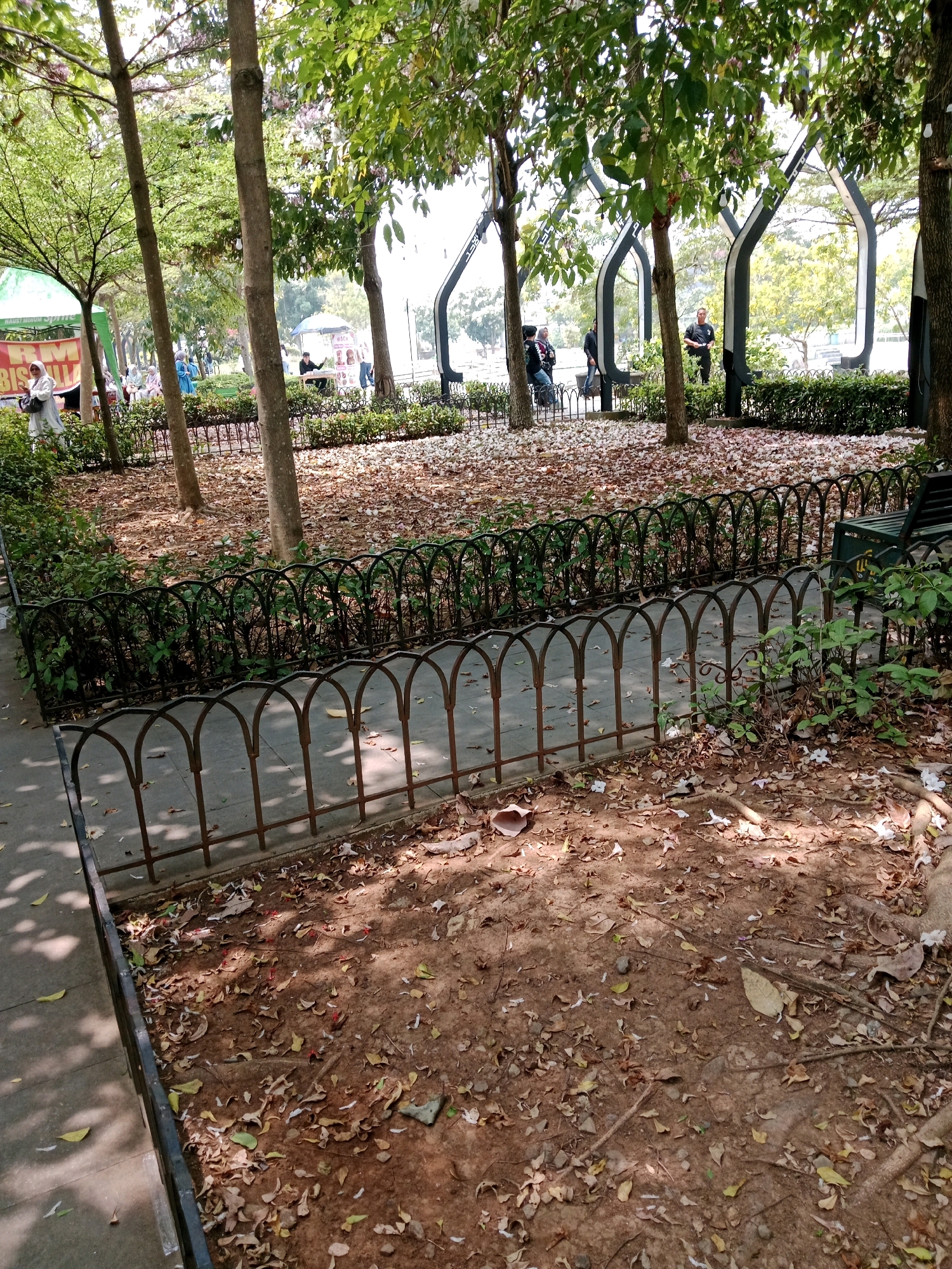 A park scene in Cianjur, Indonesia, shows a paved walkway adjacent to a dirt ground covered in fallen leaves and scattered white petals. A low, dark metal fence with arched tops separates the walkway from a patch of green bushes and trees. Beyond the bushes, more trees with sparse canopies cast dappled shadows. In the background, several people are visible in a brighter, more open area. Some are standing near stalls or awnings, while others are seated. Architectural elements, possibly part of a pavilion or shelter, with curved, pointed frames are visible among the trees. The overall atmosphere suggests a public outdoor space with natural elements and some human activity.