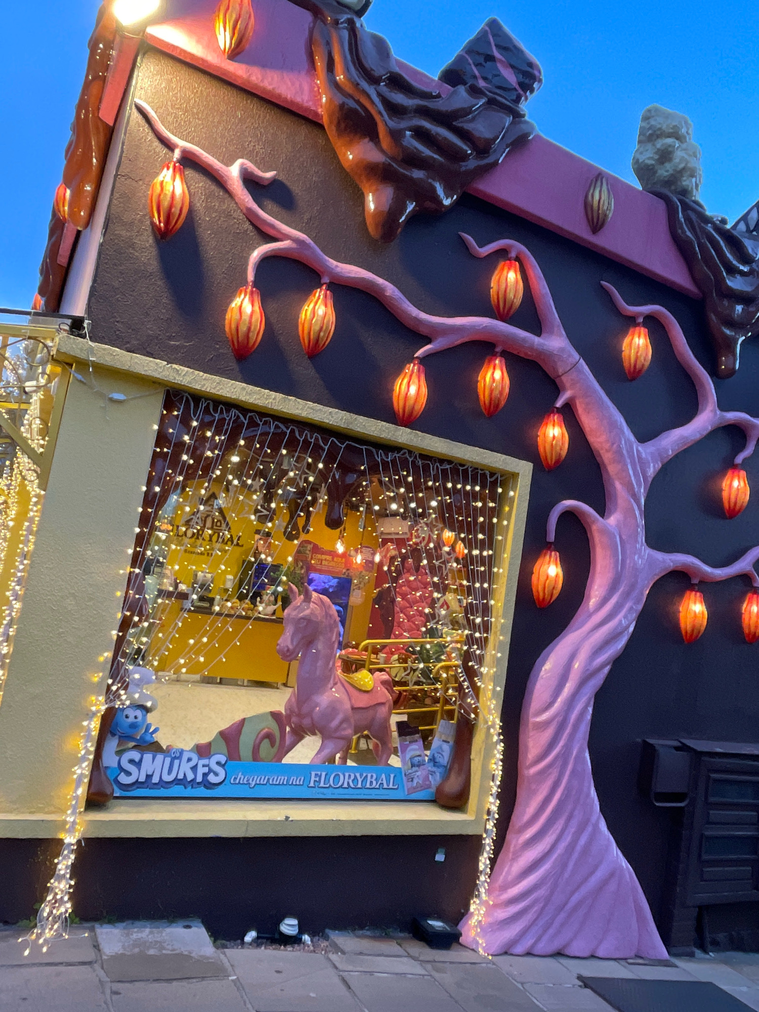 The image captures the exterior of a themed establishment, likely a shop or attraction, in Gramado, Brazil. The building features a whimsical design with chocolate-like embellishments dripping from the roof and a large, stylized pink tree with numerous illuminated fruit-shaped lamps. A yellow-framed window displays a pink rocking horse and signage for "SMURFS." The window is adorned with a dense curtain of small, white fairy lights, and similar lights also frame the left side of the building. The background shows a dark wall and hints of a dark mailbox or cabinet on the right. The ground is paved with flagstones. The overall scene suggests a festive and playful atmosphere, characteristic of tourist attractions in Gramado.