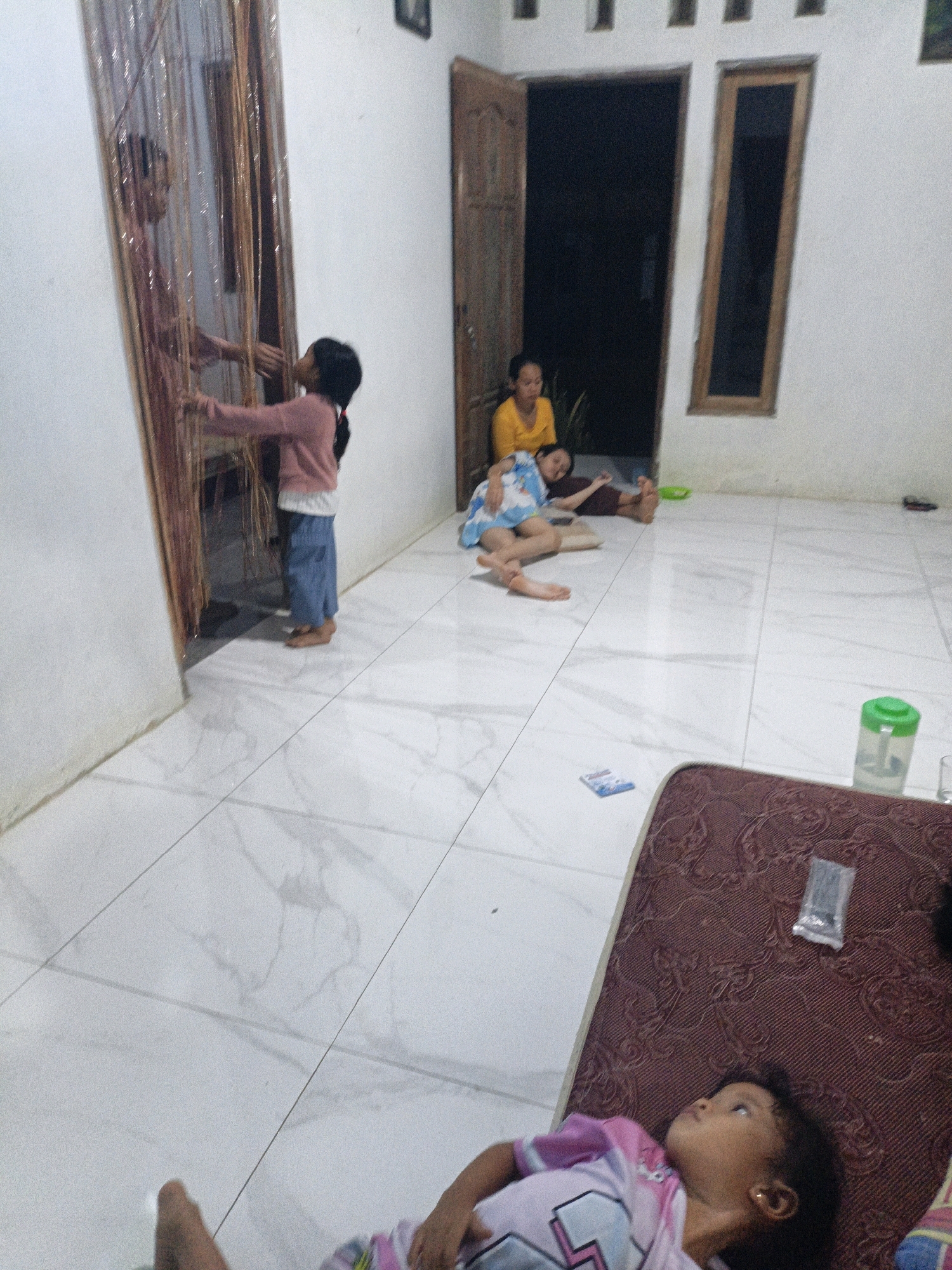 In Suwawal Timur, Indonesia, a young girl with dark hair and a pink shirt stands near a beaded curtain, her arms extended as if reaching for a reflection. The floor is tiled in a white marble pattern. Further into the room, two other individuals are seated on the floor; one woman in a yellow shirt leans back, while another person lies on the floor wearing a blue and white patterned garment. Near the seated individuals, a small green container with a white lid is visible. In the foreground, another young girl with dark hair lies on a dark maroon mattress, wearing a pink and purple t-shirt, looking upwards. The room appears to be indoors with white walls and two doorways.