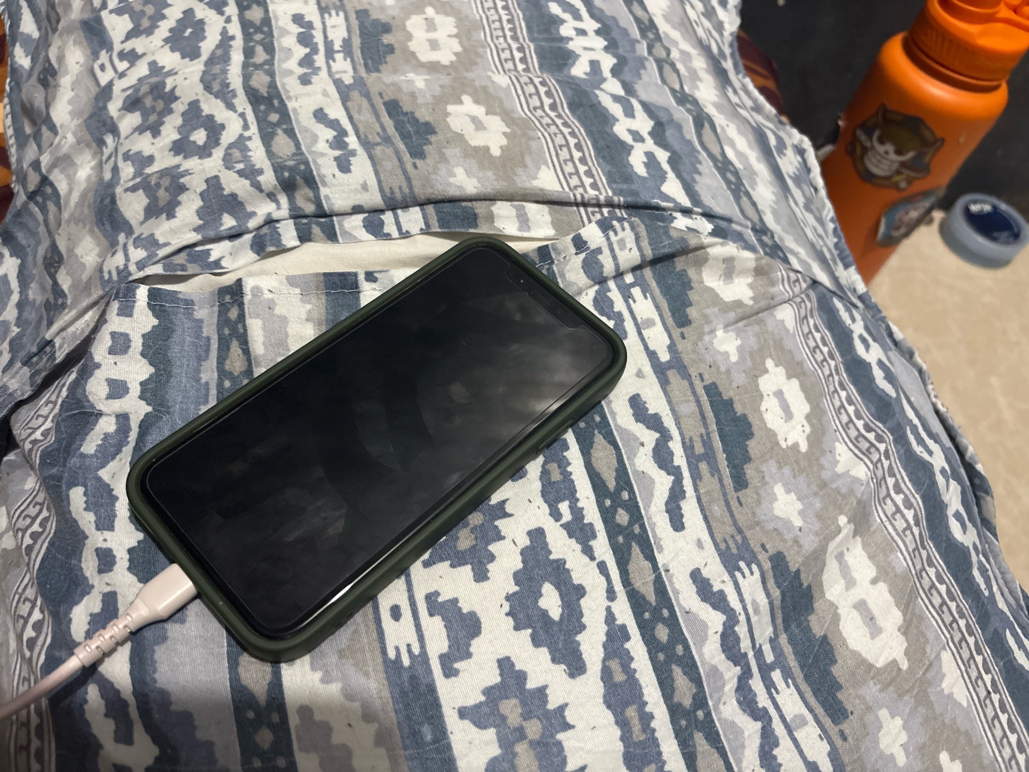 A smartphone, encased in a dark green protective cover, is placed diagonally on a patterned fabric. A light-colored charging cable is connected to the bottom of the phone. The fabric exhibits a repeating geometric design in shades of blue, gray, and white. In the background, to the right of the phone, an orange water bottle with stickers is visible, alongside a small, dark-colored circular object. The lighting suggests an indoor setting, possibly a bedroom or living area. This scene is consistent with everyday life in Malabon, Philippines.