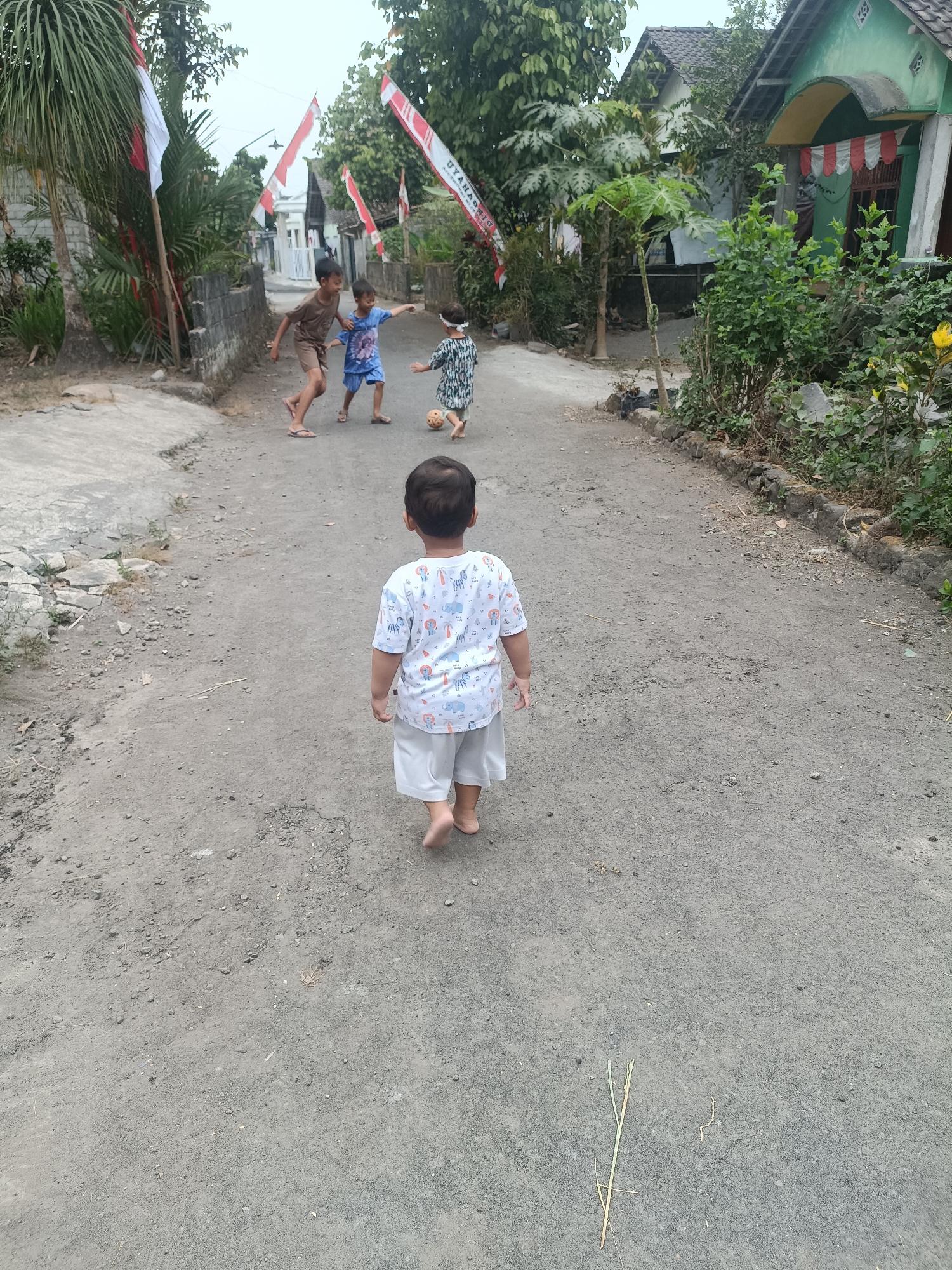 In Sumberadi, Indonesia, three young boys are playing on a dusty village road, adorned with Indonesian flags. In the foreground, a small boy wearing a white t-shirt with a print of animals and light-colored shorts walks away from the camera. Further down the road, two older boys are engaged in play. One boy, in a brown t-shirt and shorts, appears to be kicking a small ball. Another boy, in a blue t-shirt and shorts, is reaching out towards the ball. The road is unpaved and flanked by vegetation, including palm trees and various bushes. Houses with traditional Indonesian architecture are visible in the background. The sky is overcast.