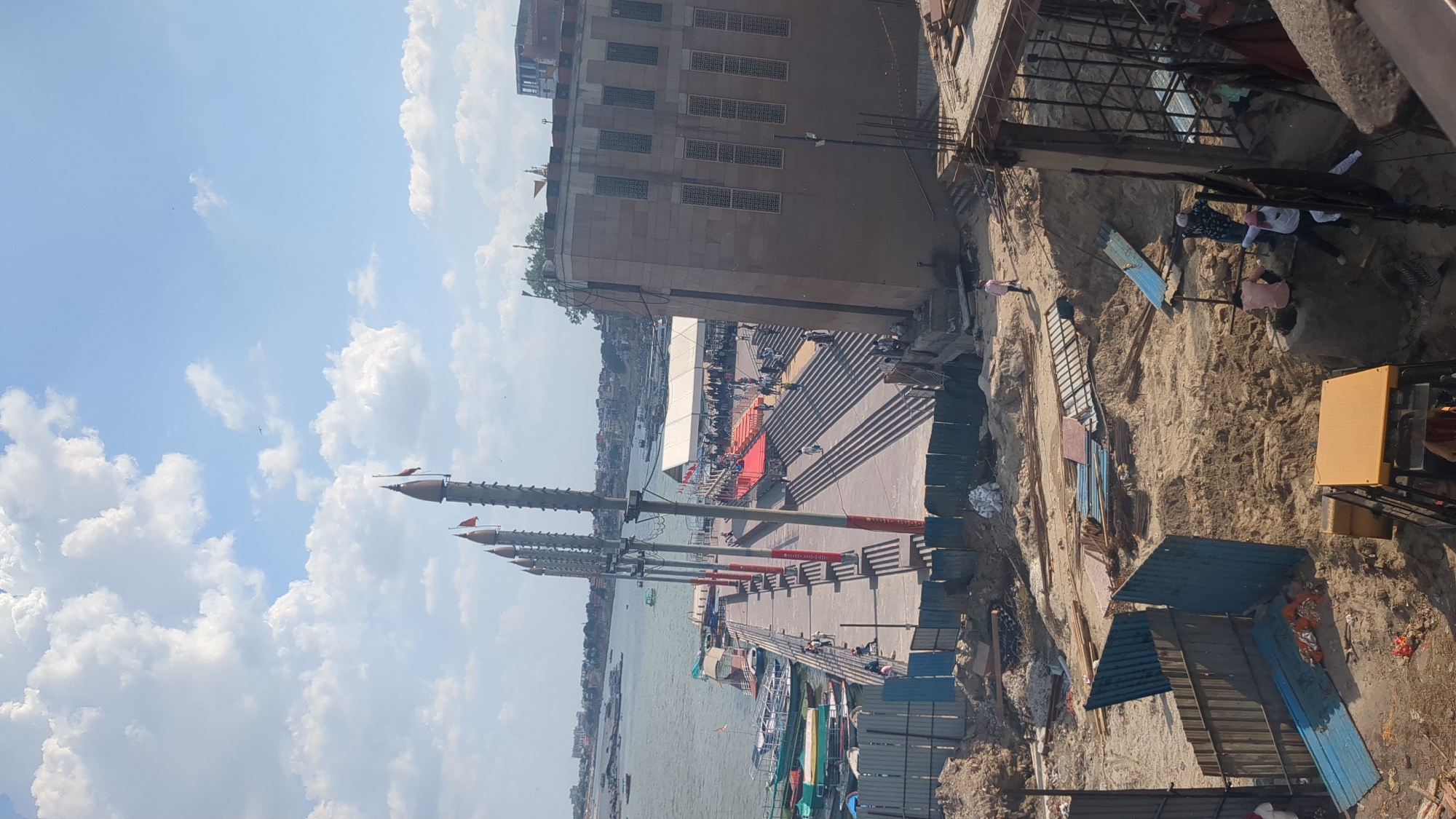 The image shows a construction site and a waterfront area in Varanasi, India. In the foreground, there is a sandy construction area with various materials, including metal sheets, rebar, and a yellow construction vehicle. Several construction workers are visible, some engaged in work and others standing or walking.
The middle ground features a promenade along the Ganges River. Several boats are docked at the waterfront. A long, elevated walkway extends from the riverbank, lined with tall poles topped with finials and flags. This walkway leads to a wide set of stairs, partially covered by a red carpet, and a large white tent structure. Numerous people are gathered in this area, suggesting an event or gathering.
In the background, across the river, the city of Varanasi is visible with its characteristic architecture and buildings. The sky is partly cloudy, with a clear blue sky dominating the upper portion of the image. The overall scene depicts a blend of ongoing construction and public activity in a significant Indian city.