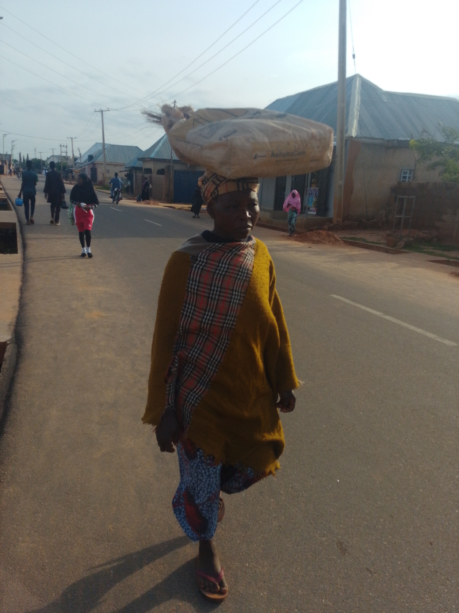 A medium shot captures a woman carrying a large sack of material on her head as she walks on an asphalt road in a town. The woman is wearing a mustard-yellow sweater over a patterned skirt, and a plaid scarf around her shoulders. She is also wearing simple flip-flops. The sack appears to be made of rough material and is strapped across the woman's head. In the background, several other people are walking on the road, and some buildings with corrugated metal roofs are visible. The sky is overcast. The scene is lit by natural light.