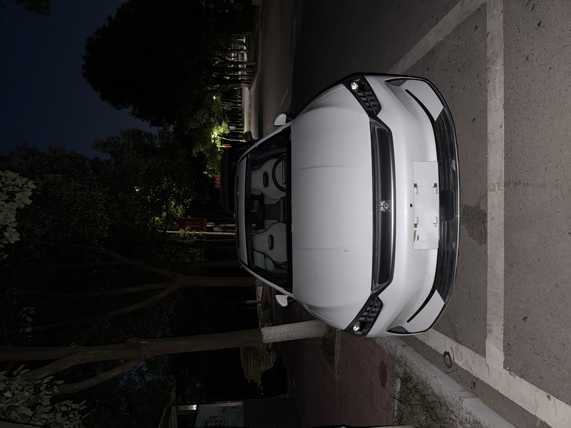 A white car is parked on the side of a road at night in Jiangjin, China. The car is a sedan with a sleek, modern design, featuring LED headlights and a prominent grille. The interior of the car is visible, showing two white racing-style seats. The car is parked in a marked parking spot, with white lines painted on the asphalt. Trees line the side of the road, and streetlights illuminate the scene. The sky is dark, indicating it is nighttime. There are no people visible in the image. The overall impression is one of a quiet, urban street at night.