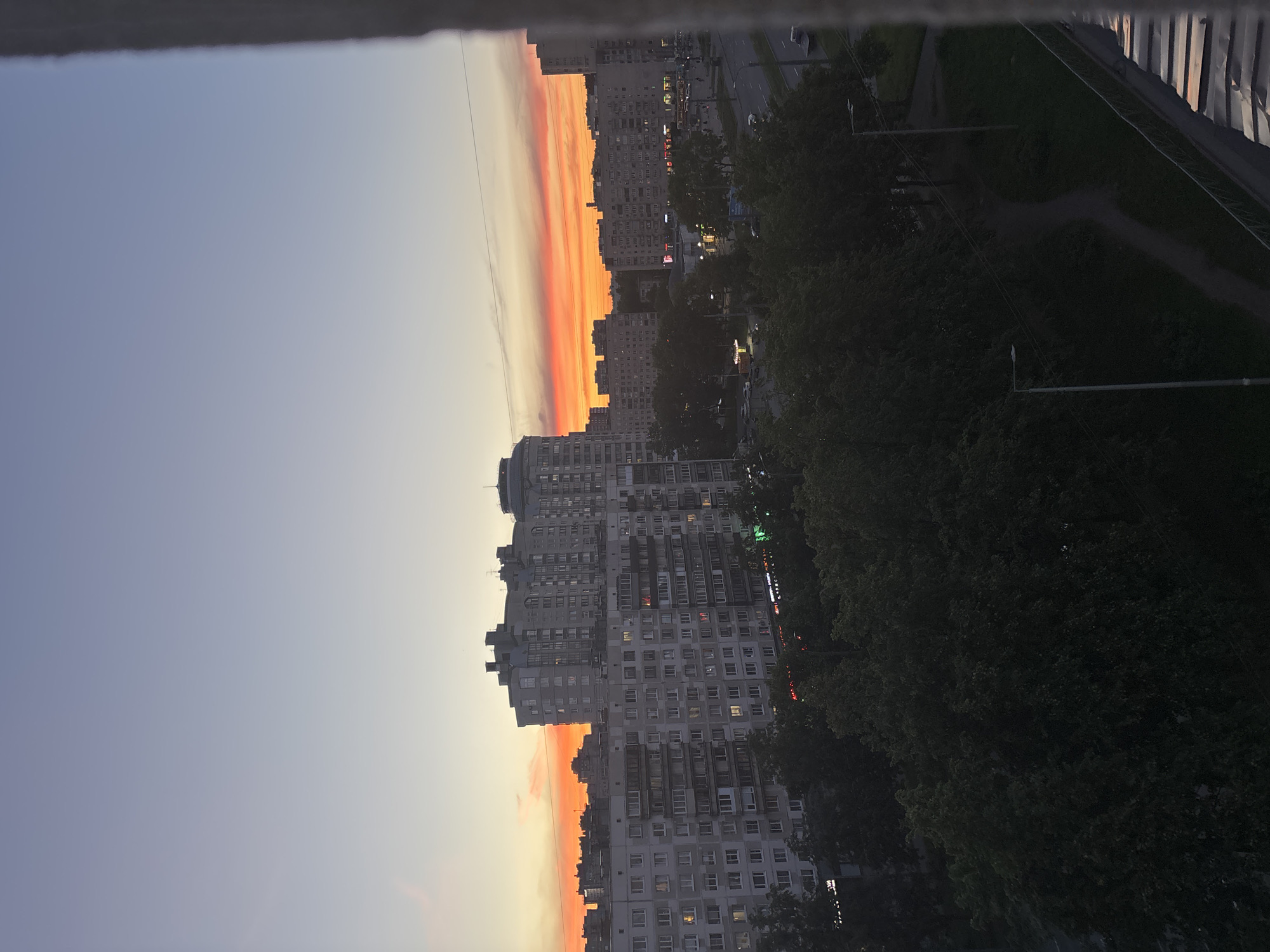 The image captures a city skyline at sunset, likely in Ladozhskoe Ozero, Russia. The sky displays a gradient of colors, transitioning from a pale blue at the top to vibrant hues of orange and red near the horizon, indicating the setting sun. Dominating the mid-ground are multi-story apartment buildings, characterized by their uniform architecture and numerous windows, some of which are illuminated. Below the buildings, a dense canopy of dark green trees lines the streets. A road is visible, with a few vehicles present. The foreground features a portion of a building or structure with a rough, textured surface, possibly a balcony railing or window frame, along with a metallic roof structure on the right. The overall scene depicts an urban environment during twilight.