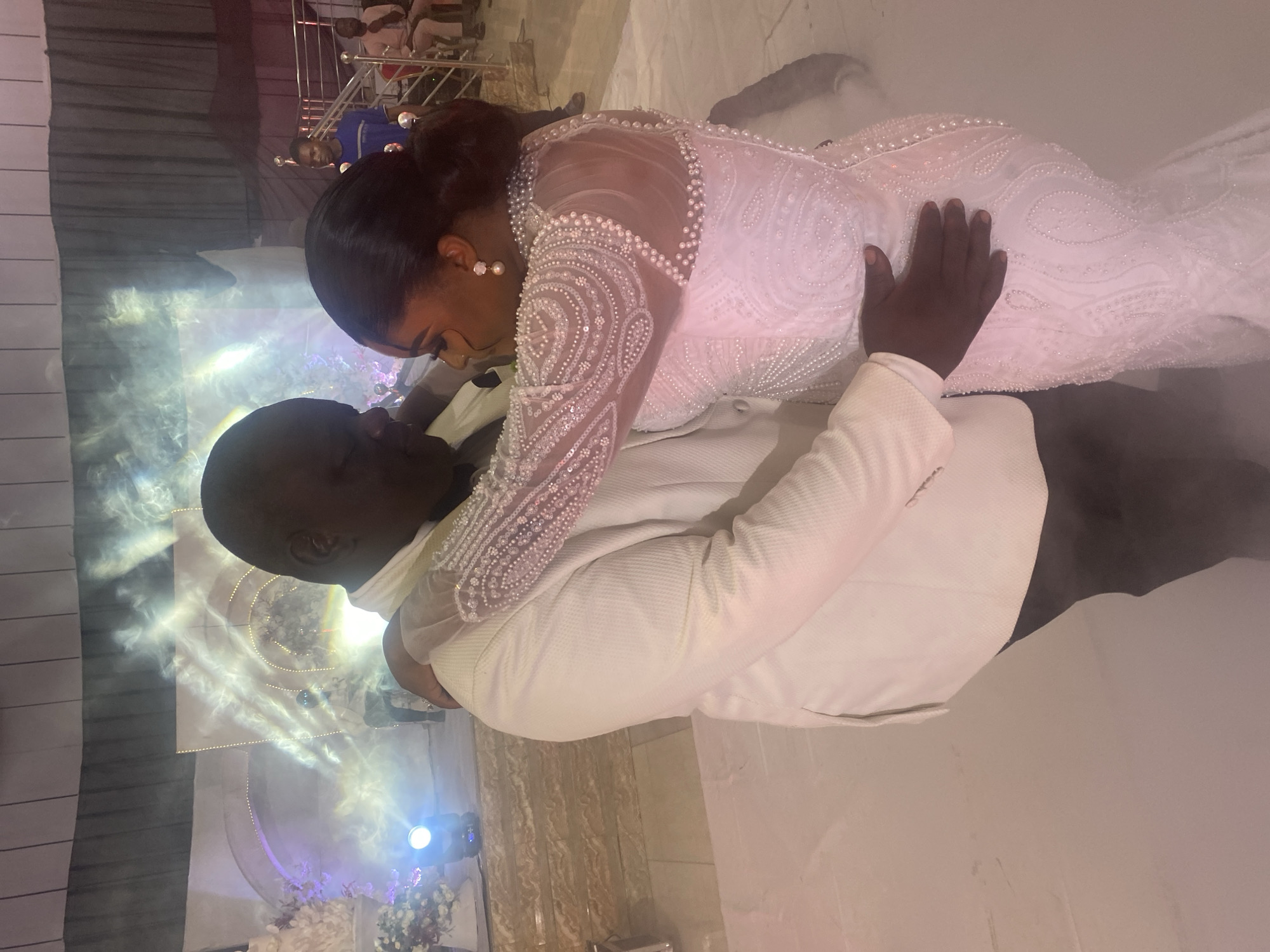 A bride in a white, pearl-embellished gown is being held by the groom, who is wearing a white suit. They are likely dancing at their wedding reception in Jos, Nigeria. A light fog covers the floor around them. There are decorations and lighting visible in the background.