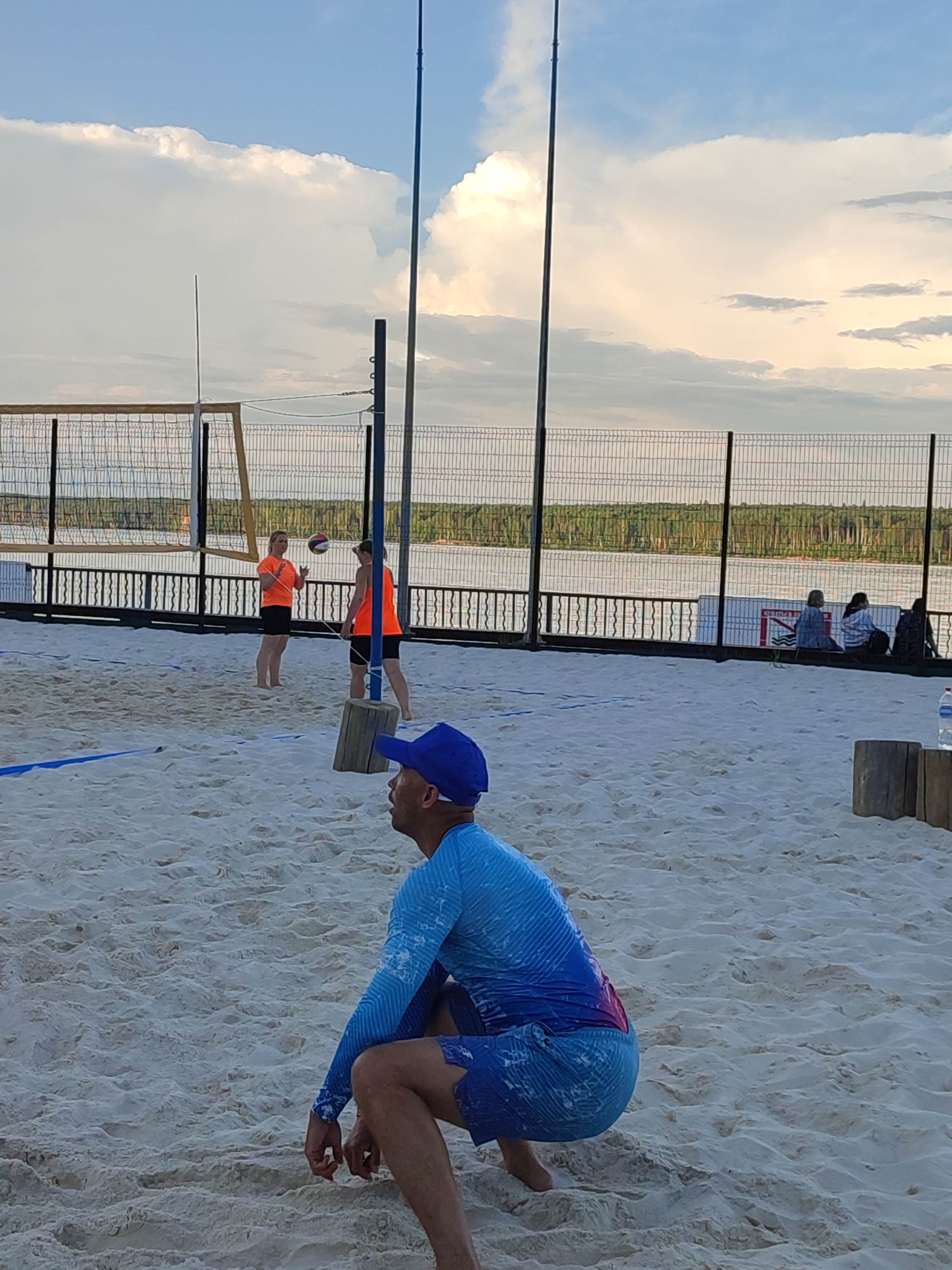 In an outdoor beach volleyball court, a man in a blue cap and long-sleeved blue shirt with blue shorts is crouched in the sand, looking to his right. The court is covered in white sand, with blue lines marking boundaries. In the background, two women in orange tops and black shorts are playing volleyball, with one about to serve. Beyond the court, a fence separates the sand from a body of water, with trees lining the far bank. Several people are sitting on a bench behind the fence, watching the game. The sky is a pale blue with large, fluffy clouds, and tall poles with volleyball nets are visible.