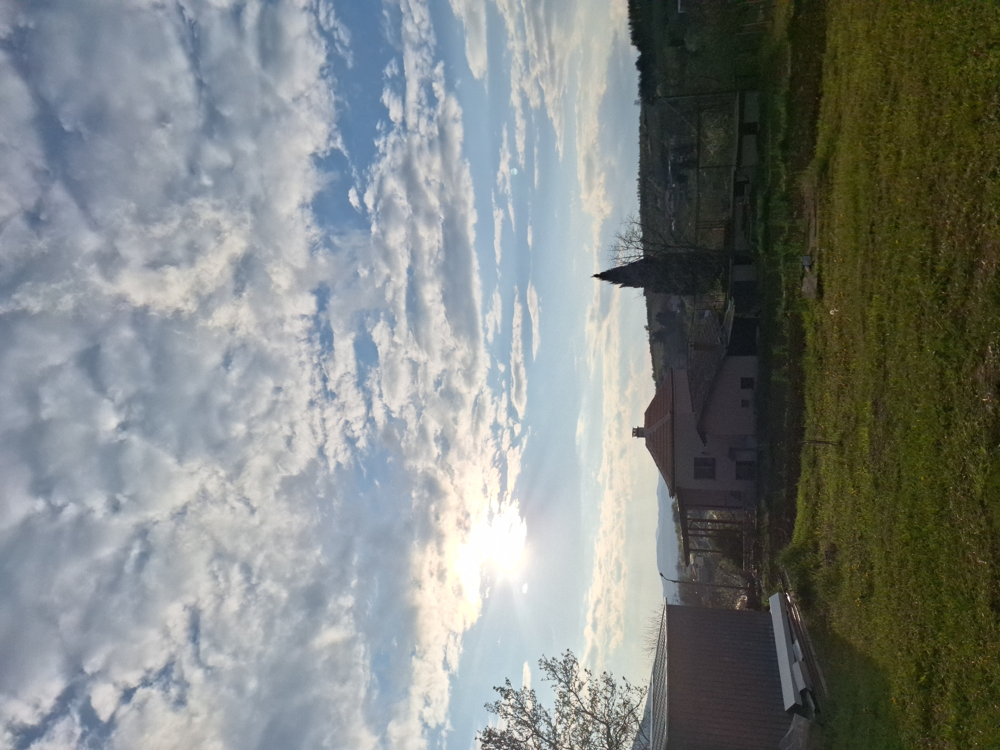 The image depicts a rural landscape under a bright, cloudy sky. The sun shines intensely, partially obscured by the clouds. In the middle ground, a building with a reddish roof and chimney is visible, along with trees and greenery. An additional structure with a gray roof appears in the foreground. A field of green grass occupies the right side of the frame. The horizon is slightly obscured by a distant tree line.