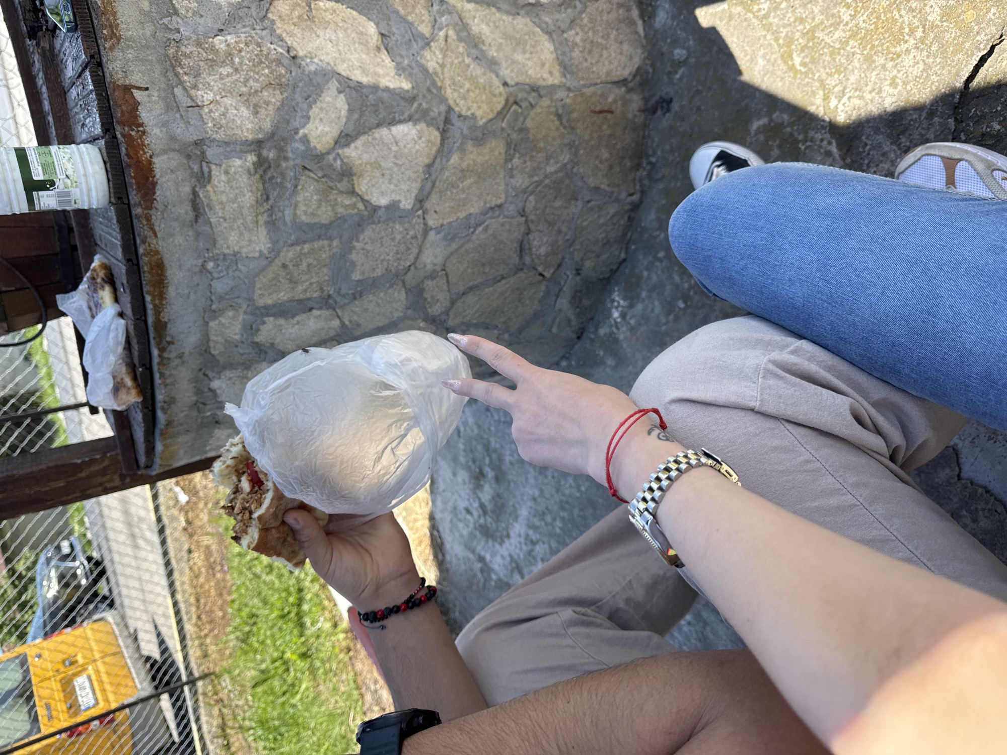 The image presents an overhead shot of two individuals, likely seated outside, sharing a meal. One person is holding a sandwich wrapped in a clear plastic bag. Their hand is adorned with a black and red beaded bracelet. On the same wrist, a black smartwatch is visible. The other person's hand is making a peace sign, and they have a red string bracelet on their wrist. A silver-colored watch is on their forearm. The individuals are wearing casual clothing, jeans and pants. The setting appears to be a roadside in Probuda, Bulgaria, indicated by the presence of a fence, a yellow van, and greenery in the background. The van's license plate reads TX-01-89-CT, and the surrounding buildings appear residential. The surface they are sitting on is made of stone.