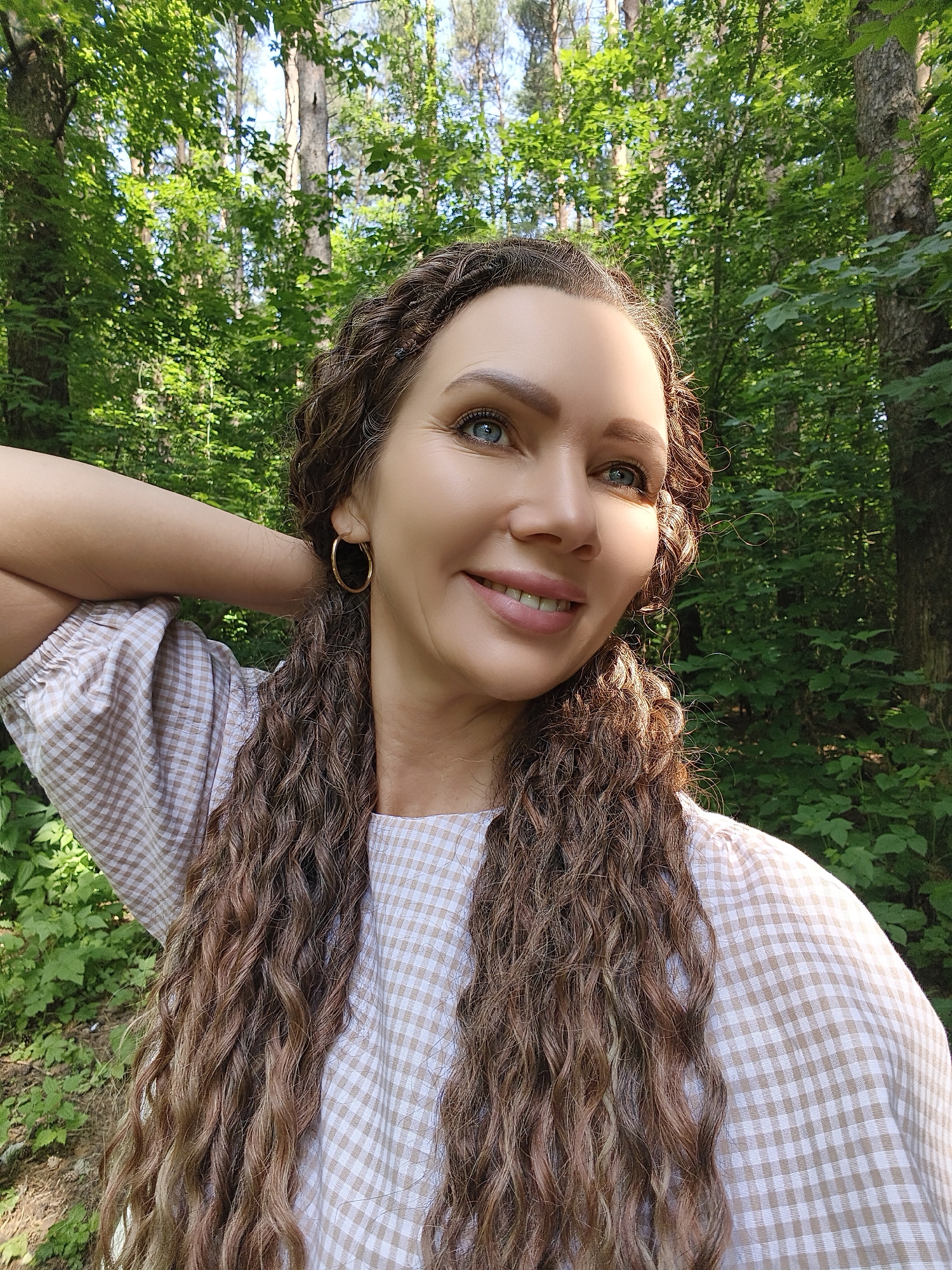 A medium shot shows a woman with long, wavy brown hair and blue eyes smiling in a forest. She is wearing a beige and white checkered dress and gold hoop earrings. Her right arm is raised behind her head, and her left hand is resting on her arm. Sunlight is casting shadows on her face, highlighting her cheekbones and the bridge of her nose. The background consists of lush green trees and foliage.