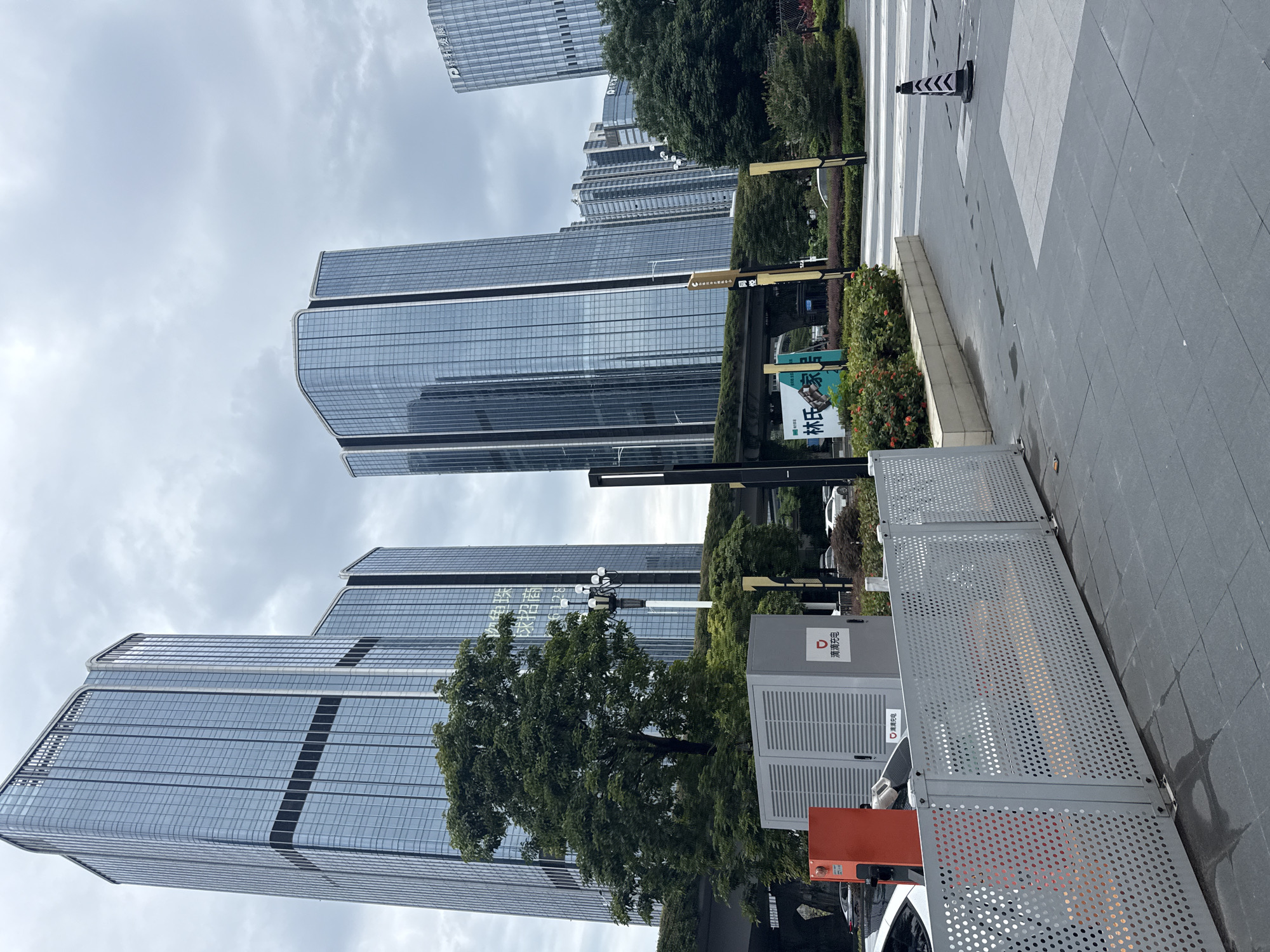 The image shows a street scene in Huangpu District, China, with modern high-rise buildings under a cloudy sky. In the foreground, a gray perforated metal fence lines a paved walkway. To the left of the fence is an orange charging station and a gray electrical enclosure with a red logo and Chinese characters that translate to "Charging Pile." Behind the fence, a white car is parked, and a street crossing with a traffic cone is visible. In the mid-ground, lush green trees and bushes add color to the urban landscape. The background features several tall, glass-fronted office buildings, reflecting the overcast sky. A green bridge or elevated walkway with vegetation on top spans across the scene, connecting some of the buildings. The overall impression is one of a contemporary business district with a focus on infrastructure and greenery.