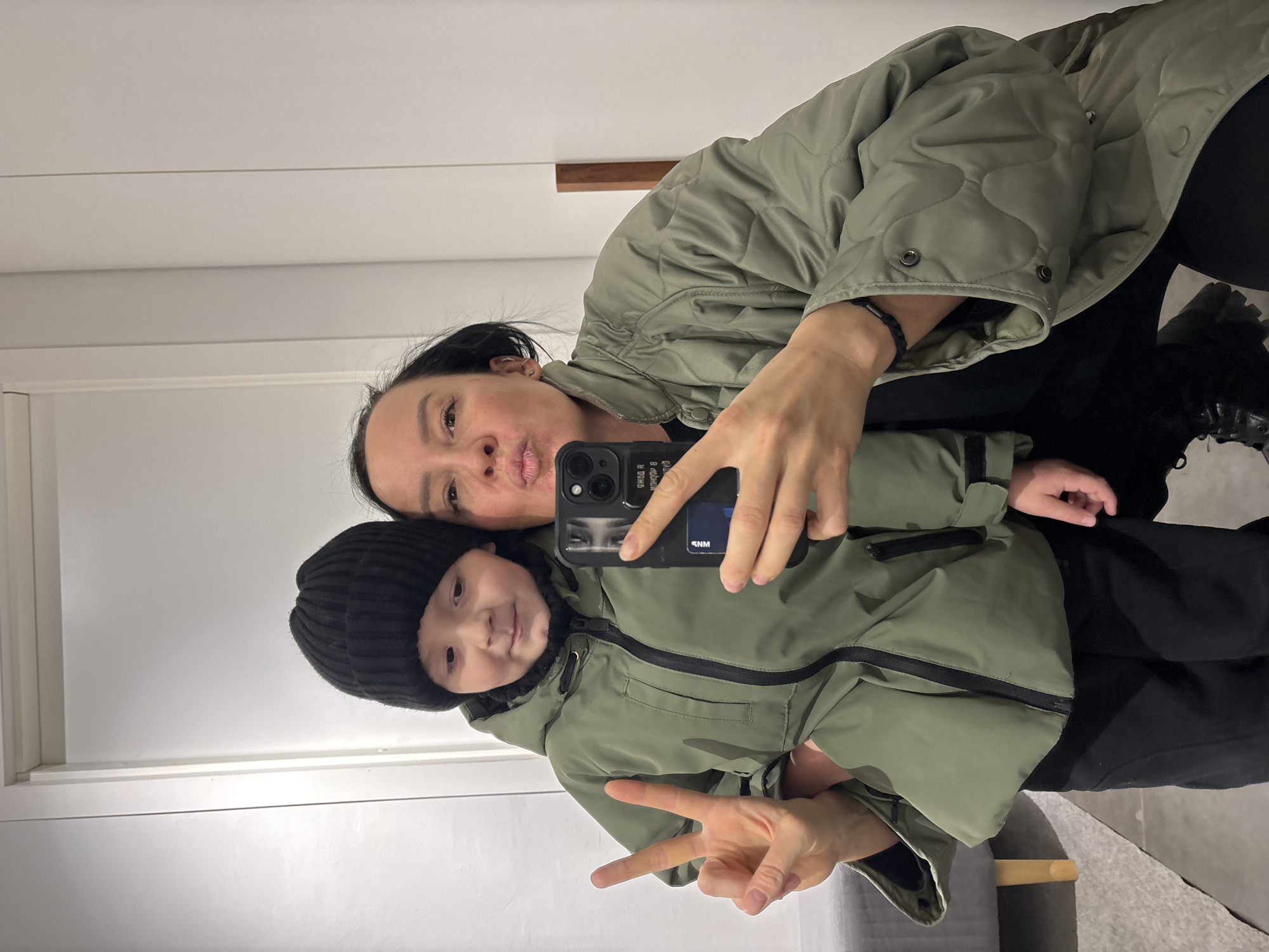An adult female and a young child pose for a selfie in an indoor setting in Chaikovsky, Russia. The image is taken from a slightly high angle, showing them both from the chest up.
The adult, with dark hair, wears a light olive green quilted jacket over a dark top. She has a dark bracelet on her left wrist and is making a playful puckering or kissing face towards the camera. She holds a black smartphone with multiple camera lenses and a black case in her right hand, taking the selfie. Reflections on the phone case show what appear to be eyes and some illegible text, possibly "snm".
The child, positioned in front of the adult, wears a black beanie and a light olive green puffer jacket with black zippers. The child has a gentle smile and holds up a peace sign with their left hand. Both individuals appear to be in good spirits.
The background consists of plain, light-colored walls with a subtle horizontal line across them, suggesting architectural detail or paneling. A narrow wooden element is visible on the upper right side. Part of a grey surface, possibly a bench or floor, is visible at the bottom of the frame. The jackets and beanie suggest a cool climate, though the photo is taken indoors.