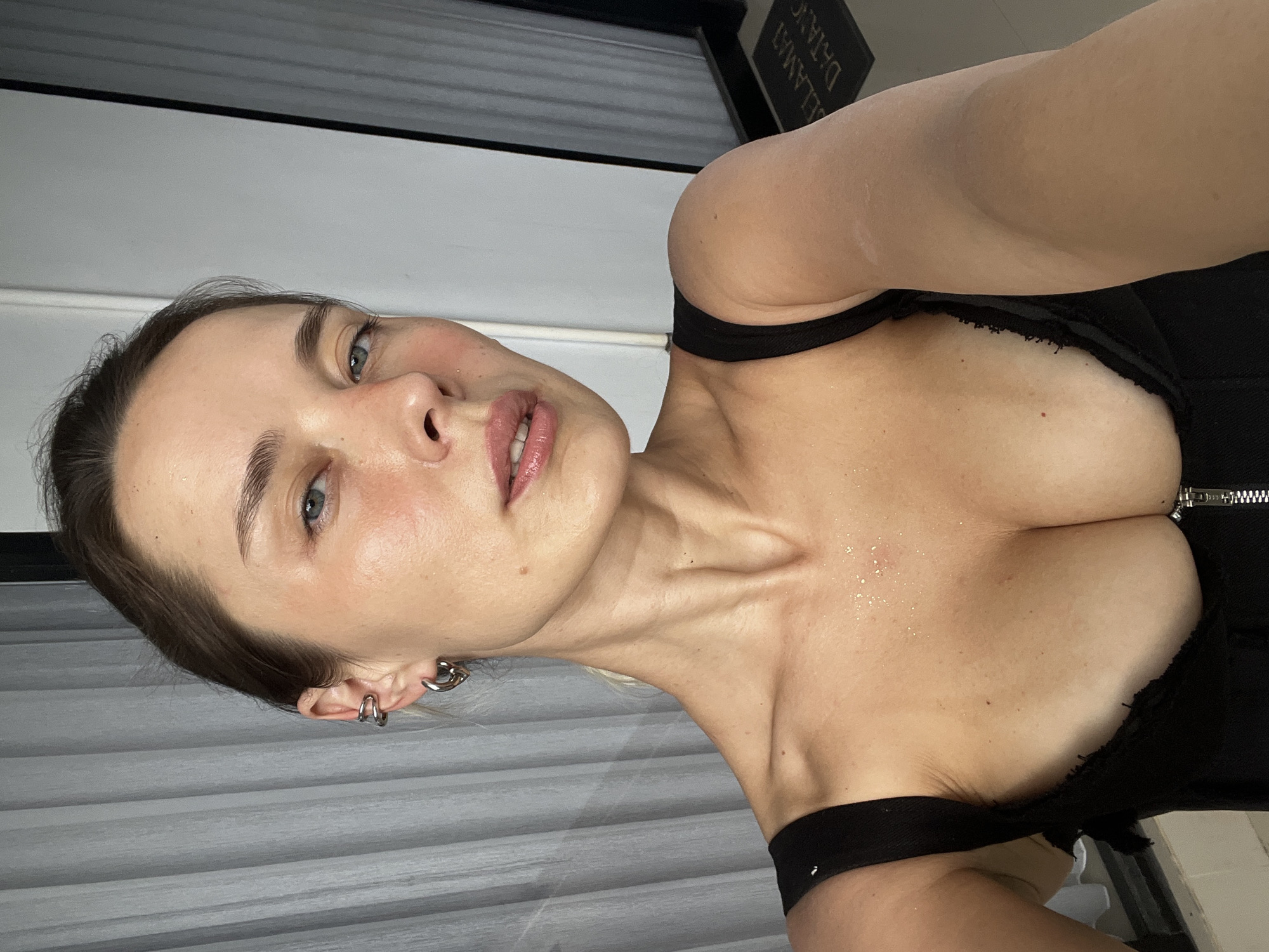 A woman is taking a selfie in front of a sheer gray curtain and a glass door. She has blonde hair pulled back into a bun, blue eyes, and is wearing a black, low-cut top with a zipper. She has multiple silver earrings in her left ear. The background suggests an indoor setting with a glimpse of a doormat with the words "SELAMAT DATANG" visible. The lighting is natural and appears to be coming from the side.