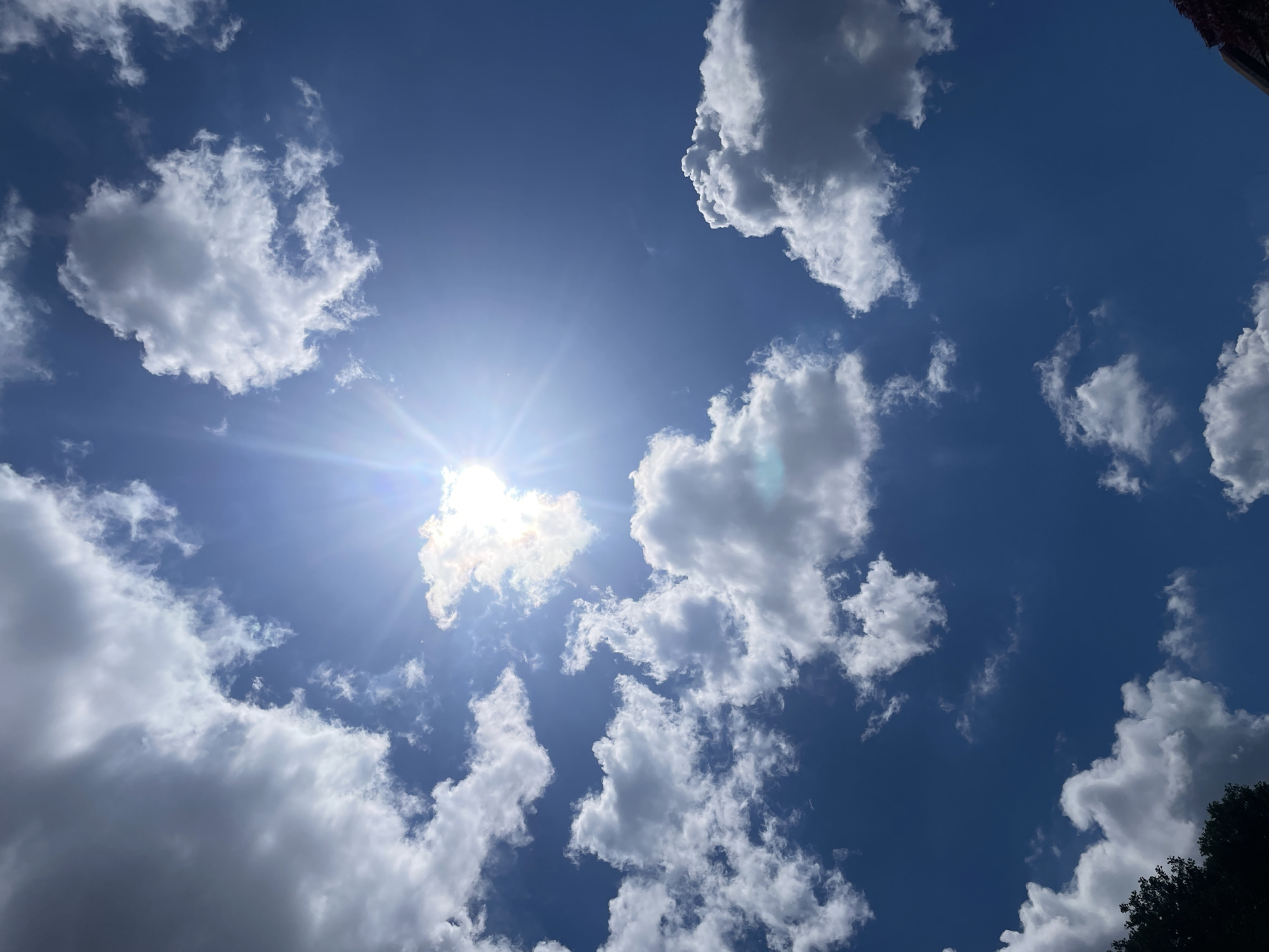 This is a vertical, eye-level, medium shot of a bright blue sky with scattered white clouds. The sun is prominently visible in the upper center of the image, with strong rays of light emanating from it. Some of the clouds are catching the sunlight, appearing bright white, while others are darker and appear to be in shadow. The lower left corner of the image shows the dark silhouette of tree branches, and in the lower right corner, a dark, angled structure, possibly a building or roofline, is partially visible. The overall impression is of a sunny, partly cloudy day.