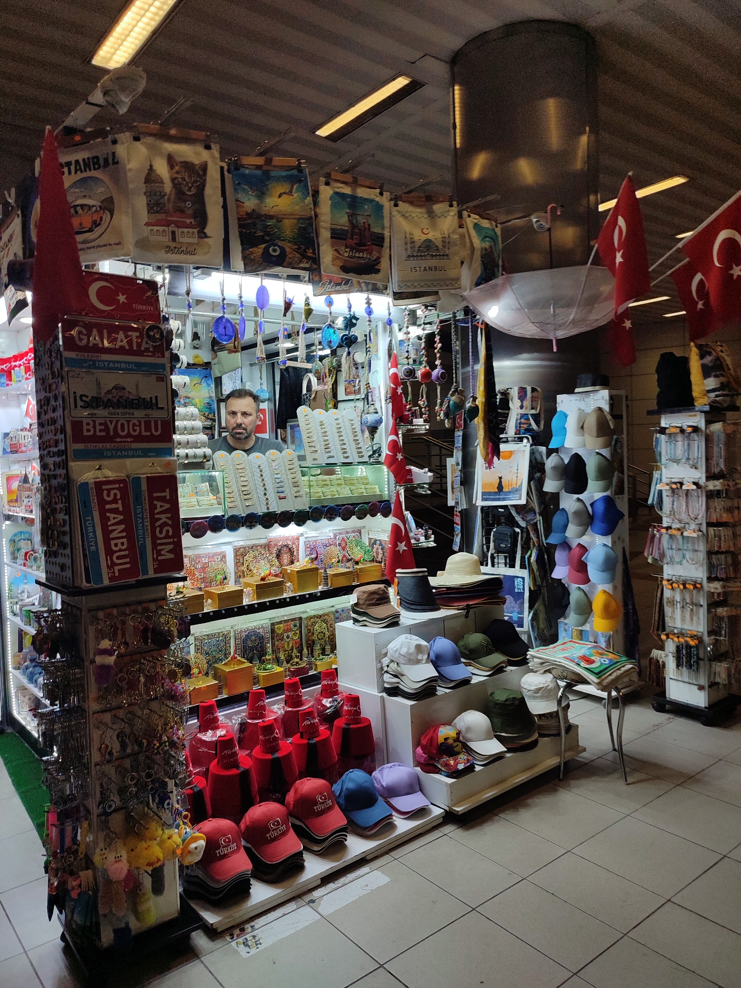 This image depicts a souvenir shop, likely located in the Beyoğlu district of Istanbul, Turkey, given the prevalent imagery of Istanbul, Galata, and Taksim. A man with dark hair and a beard, wearing a grey t-shirt, stands behind a counter, appearing to be a vendor. The shop is filled with various tourist merchandise, including hats, keychains, Turkish flags, magnets, decorative plates, and textiles featuring local landmarks and symbols like the Hagia Sophia and cats. The display units are well-stocked and organized, showcasing a wide array of souvenirs intended for tourists. The lighting appears to be artificial, emanating from fluorescent tubes overhead, casting a warm glow on the merchandise. The floor is tiled with beige and light grey squares. The overall impression is that of a bustling market stall designed to attract and cater to international visitors.