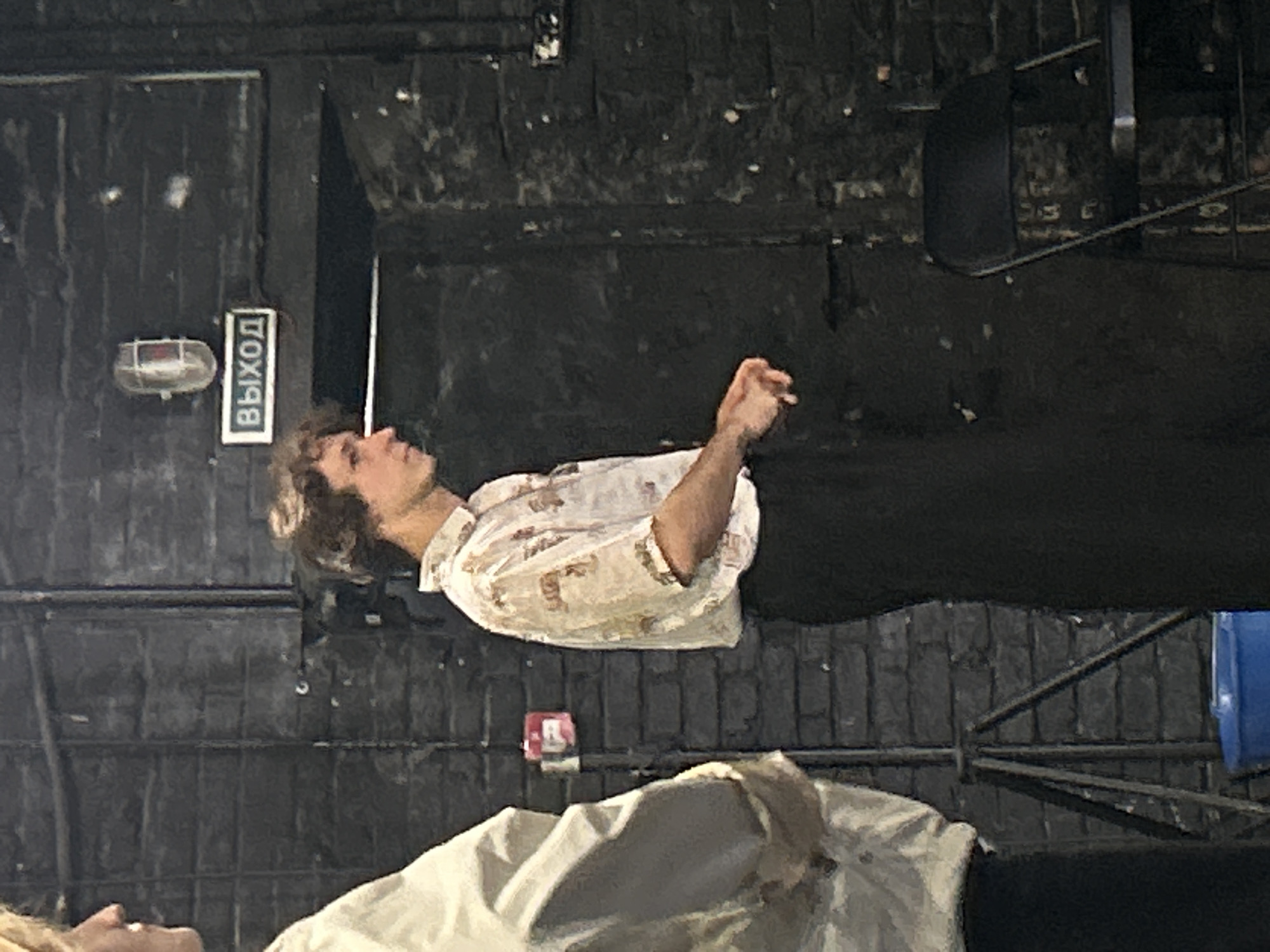 In the city of Unknown, Unknown, a young man with curly brown hair stands in profile, facing right. He is wearing a short-sleeved, collared shirt with a patterned print and dark trousers. His hands are clasped in front of him. To his left, another person, only partially visible from the shoulders up and wearing a light-colored garment, is also present.
The background consists of a dark brick wall. Mounted on the wall is a rectangular sign with the word "ВЫХОД" (which translates to "EXIT" in Russian) written in white letters on a black background, accompanied by a light fixture above it. A red square object, possibly an alarm or a button, is also attached to the wall.
In the foreground, a dark-colored tripod stands on the left, and a blue bucket is visible at its base. A black folding chair is positioned to the right of the scene. The overall lighting is dim, suggesting an indoor environment like a theater or a rehearsal space.