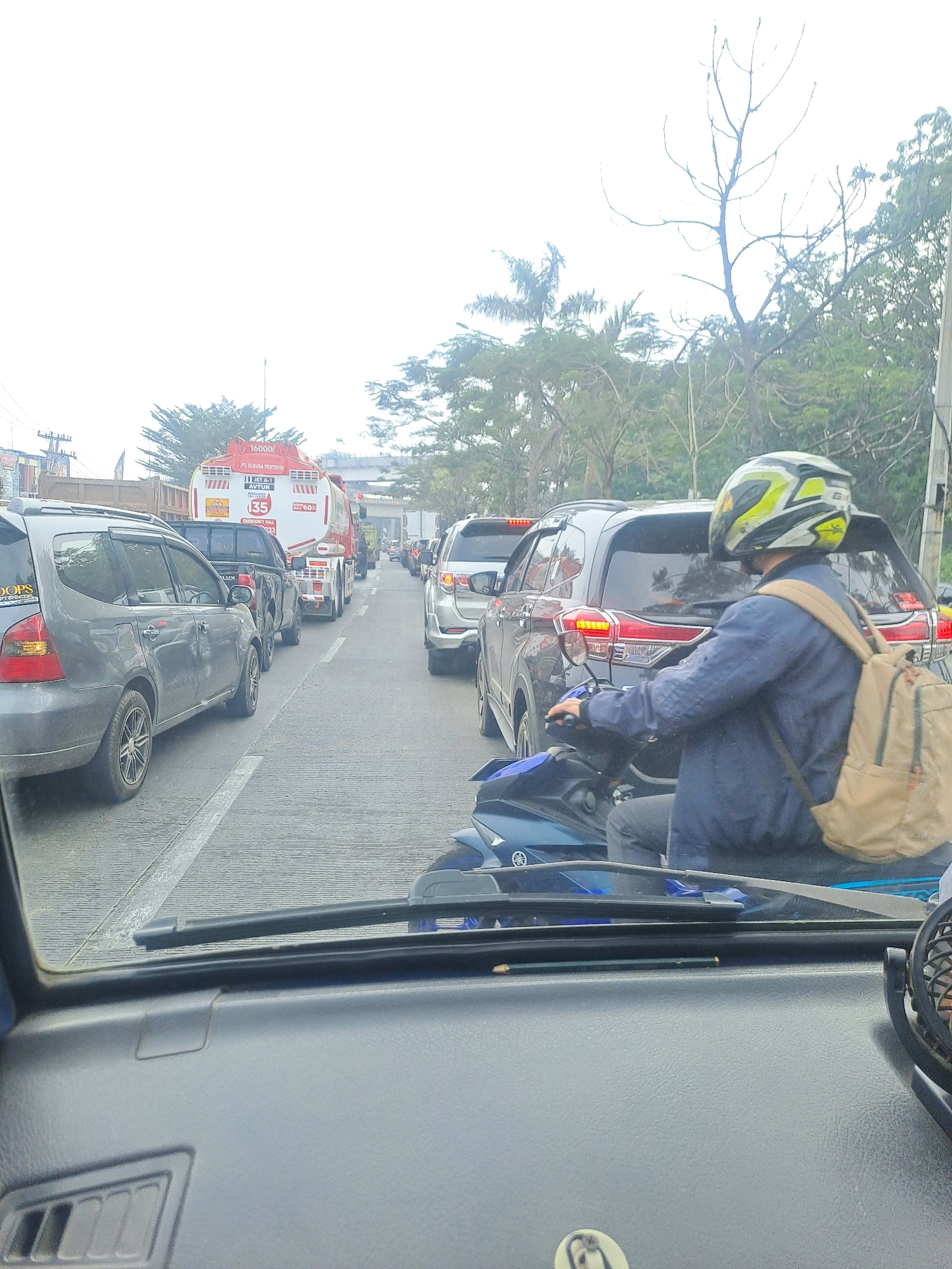 The image depicts a traffic jam on a road in Palembang, Indonesia, likely during daytime. The perspective is from inside a vehicle, looking forward through the windshield. Several cars, including a grey minivan and a black SUV, are visible, stopped in traffic. A large red and white tanker truck with the text "16000l" and "PT. ELIKURA PERTOFIN" is prominent in the middle lane. A motorcyclist wearing a helmet and a blue jacket, with a tan backpack, is in the right lane, also stopped. Trees and utility poles line the side of the road. The sky is overcast, contributing to a slightly hazy atmosphere. The road surface appears wet, suggesting recent rain.