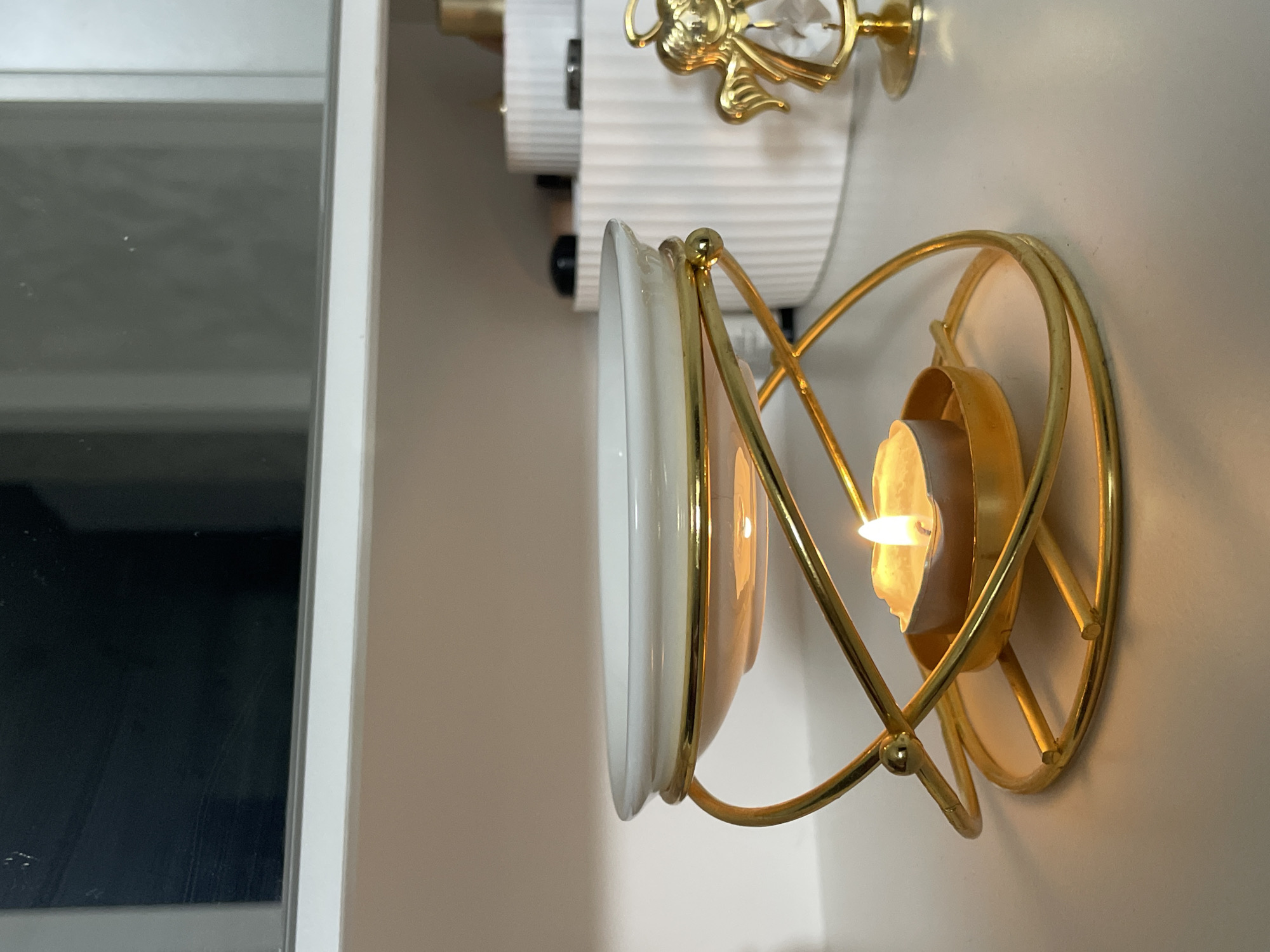A lit candle lamp is affixed to a beige wall next to a white shelf or cabinet. The lamp features a cylindrical white shade trimmed with gold, and a gold metal cage that holds the candle. The candle casts a warm glow. To the left is part of a shelf or cabinet unit, also white. The upper left of the image shows a reflection in a glass or polished surface. The specific location is not evident from the image.