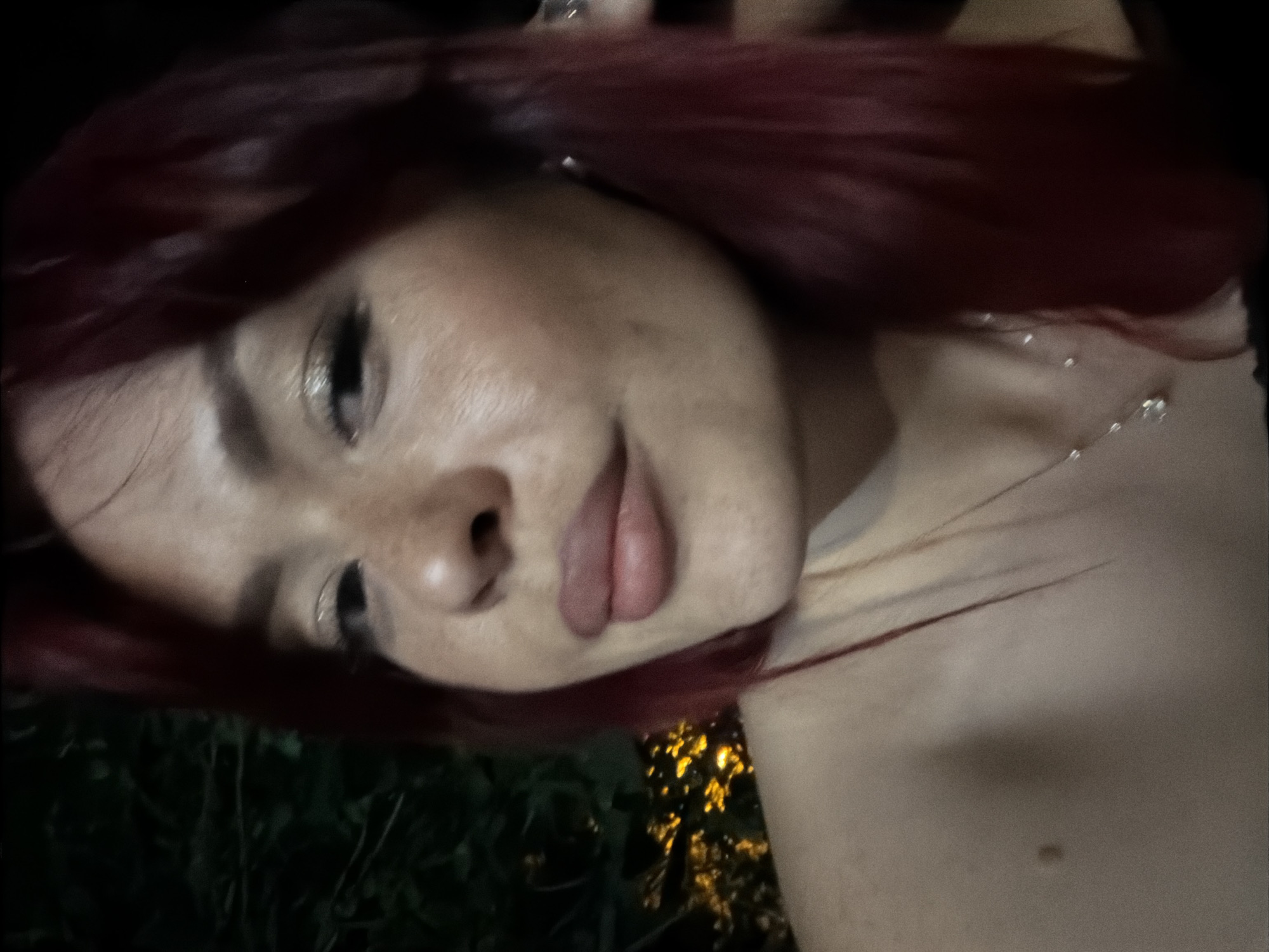 The image is a close-up selfie of a woman with dark red hair, taken at night. She is looking at the camera with a slight smile. Her makeup includes eyeshadow and lipstick. She is wearing a necklace with a pendant. The background is dark, with some out-of-focus lights visible, suggesting an outdoor setting, possibly a park or a garden. There are no discernible landmarks or features that would indicate the specific location within the city of Tolyatti, Russia.