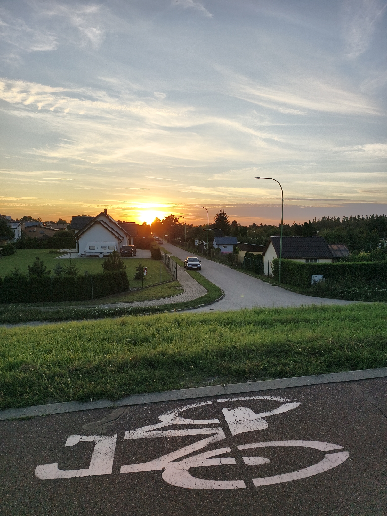 The image depicts a suburban street scene in Tczew, Poland, during a sunset. The foreground features a paved surface with a white bicycle lane marking. To the left of the bicycle lane is a sloping grassy embankment. Beyond the embankment, a residential street curves to the right, lined with houses, fences, and trees. A white camper van and a dark SUV are parked in the driveway of a house on the left. A silver sedan is parked on the street further down. A streetlamp is visible on the right side of the street. The sky is illuminated by the setting sun, casting a warm glow over the scene and creating wispy clouds. The overall atmosphere is peaceful and serene.