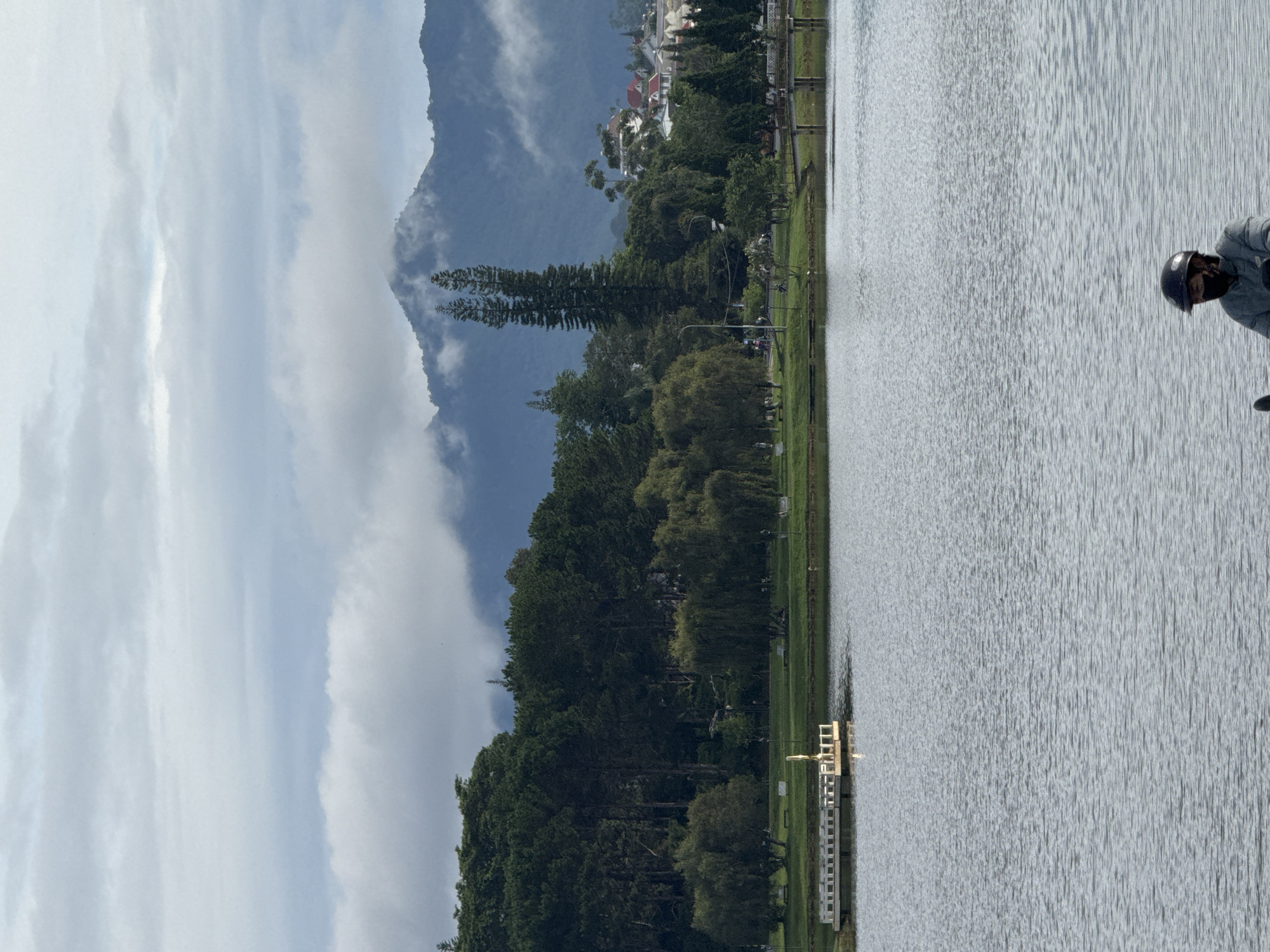 This media file depicts a serene lake scene with a person on a bicycle in the foreground, facing away from the camera. The person is wearing a dark helmet and a grey jacket, suggesting a cool or mild day. The lake itself occupies a significant portion of the right side of the image, its surface rippling with gentle waves.
Along the left bank of the lake, a lush green grassy area is visible, dotted with trees of varying heights and types. In the background, a majestic mountain range rises, partially shrouded in soft, white clouds that blend into the hazy sky. The mountain slopes are covered in dense, dark green foliage. Further in the distance, nestled among the trees and foothills, are buildings with red-tiled roofs, indicating a settlement or town.
A small, white wooden dock or pier extends from the grassy bank into the water on the left side. Several people can be seen walking or standing along the path by the lake, and some motorbikes are parked near the trees. The sky is a mix of blue and white clouds, indicating it could be late morning or early afternoon. The overall atmosphere is peaceful and picturesque, typical of a natural landscape setting in a mountainous region, likely Da Lat, Vietnam, known for its lakes, pine forests, and cool climate. No visible text is present in the image.