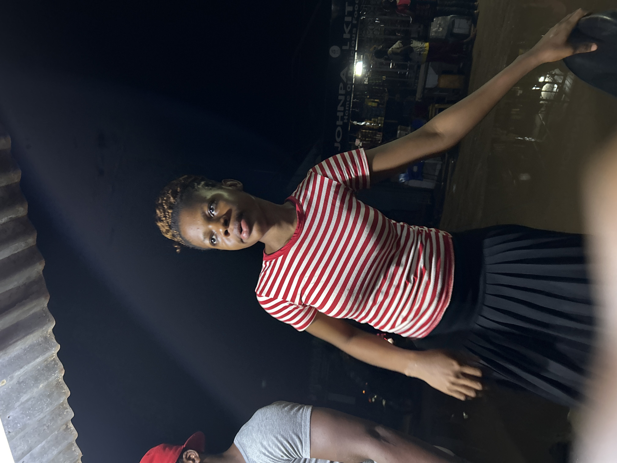 The image shows a woman wearing a red and white striped shirt and a black skirt. She is holding a black object in her raised hand. The background suggests an outdoor setting at night in Ihiagwa, Nigeria, with some indistinct structures and light sources, possibly indicating a commercial area. Another individual wearing a grey shirt and a red cap is partially visible at the bottom of the frame.