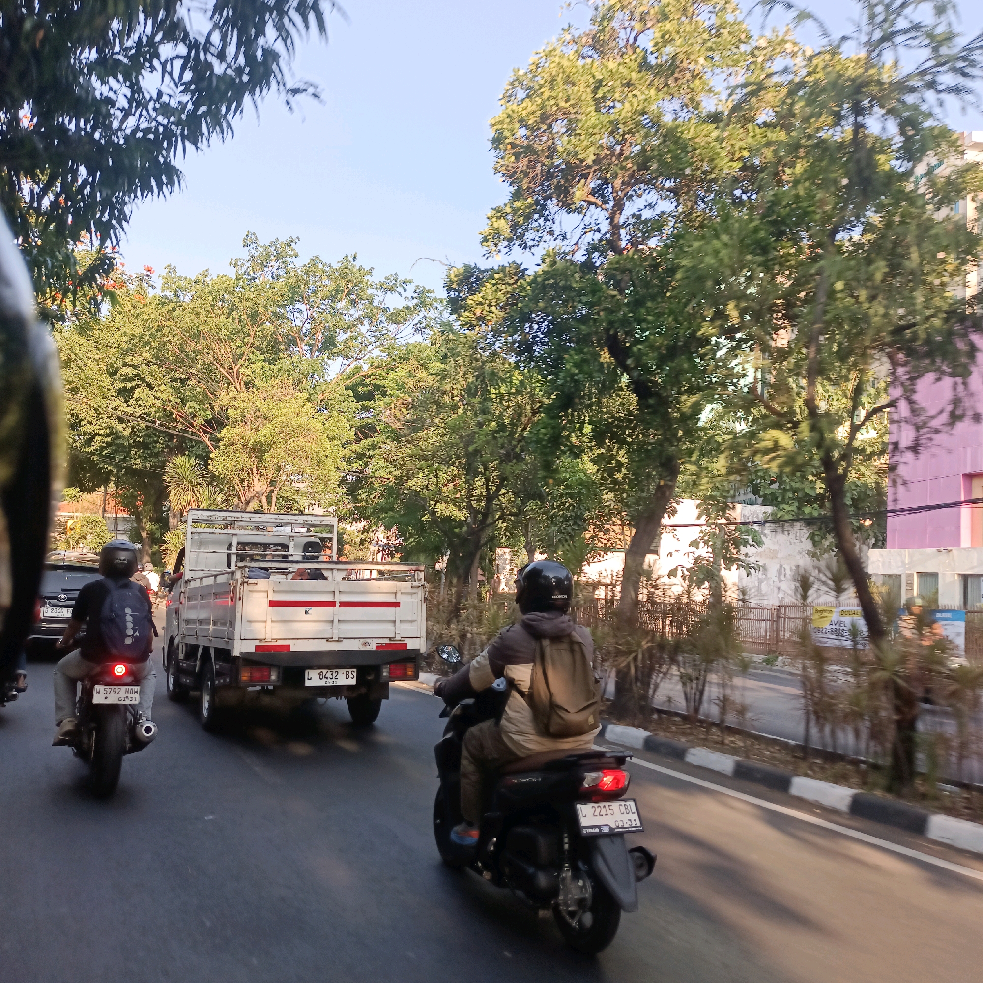 A bright, sunny daytime scene captures a busy street in Surabaya, Indonesia. The perspective is from behind a motorcycle rider, looking forward. In the immediate foreground, the handlebars and a portion of the black motorcycle are visible. Ahead, two motorcycles and a white truck are traveling on the asphalt road. The motorcycle directly in front, also viewed from behind, has a rider wearing a dark helmet and a backpack, with a license plate displaying "W 5792 NAH". To the right of this motorcycle, a white truck with a red stripe along its side is traveling, its license plate reading "L 8432 BS". Another motorcycle, this one black, with a rider also wearing a helmet and backpack, is to the right of the truck, its license plate showing "L 2215 CBL". In the background, lush green trees line the street, with a multi-story pink building visible beyond them. The sky is clear and blue. The overall impression is of a typical urban commute in a tropical climate.