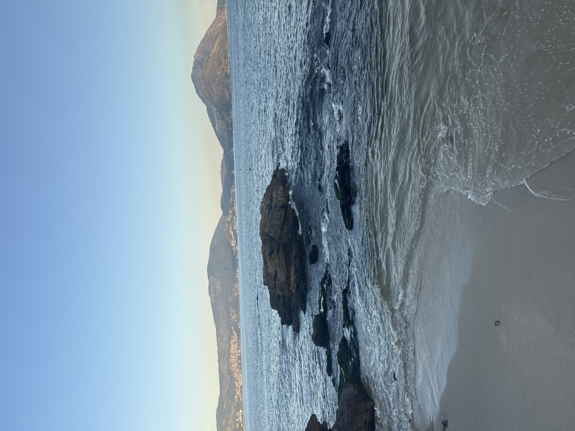 The image captures a serene view of the coast in Niterói, Brazil, likely from a beachside vantage point. The foreground features a sandy shore with gentle waves lapping at the edge, revealing some seaweed-covered rocks and larger, darker rocks emerging from the water. The middle ground is dominated by the calm, blue ocean, with a prominent cluster of dark rocks jutting out. In the background, a chain of undulating hills and mountains forms the cityscape of Niterói, dotted with buildings that are visible in the distance. The sky is a clear, pale blue, transitioning to a lighter hue near the horizon, suggesting it might be late afternoon or early morning. The overall scene evokes a peaceful, natural landscape integrated with urban elements.